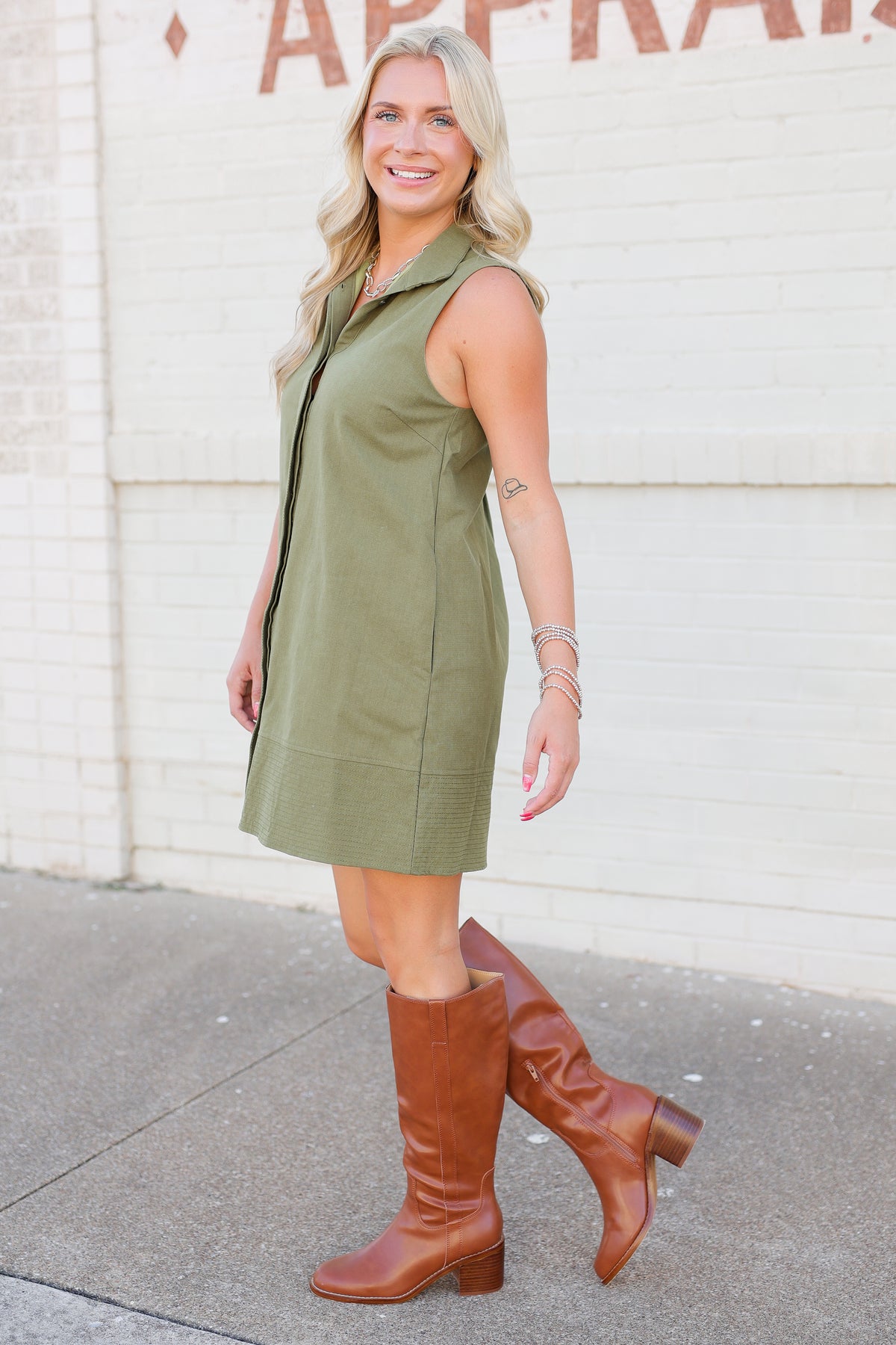 A woman wearing an olive green sleeveless collared dress and brown leather knee-high boots, standing outdoors against a white brick wall.