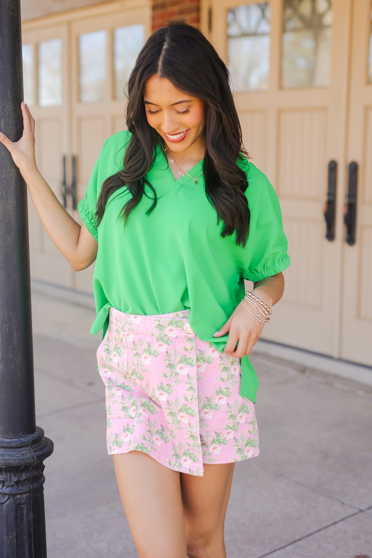 Woman wearing a green blouse and pink floral skirt standing next to a building entrance.
