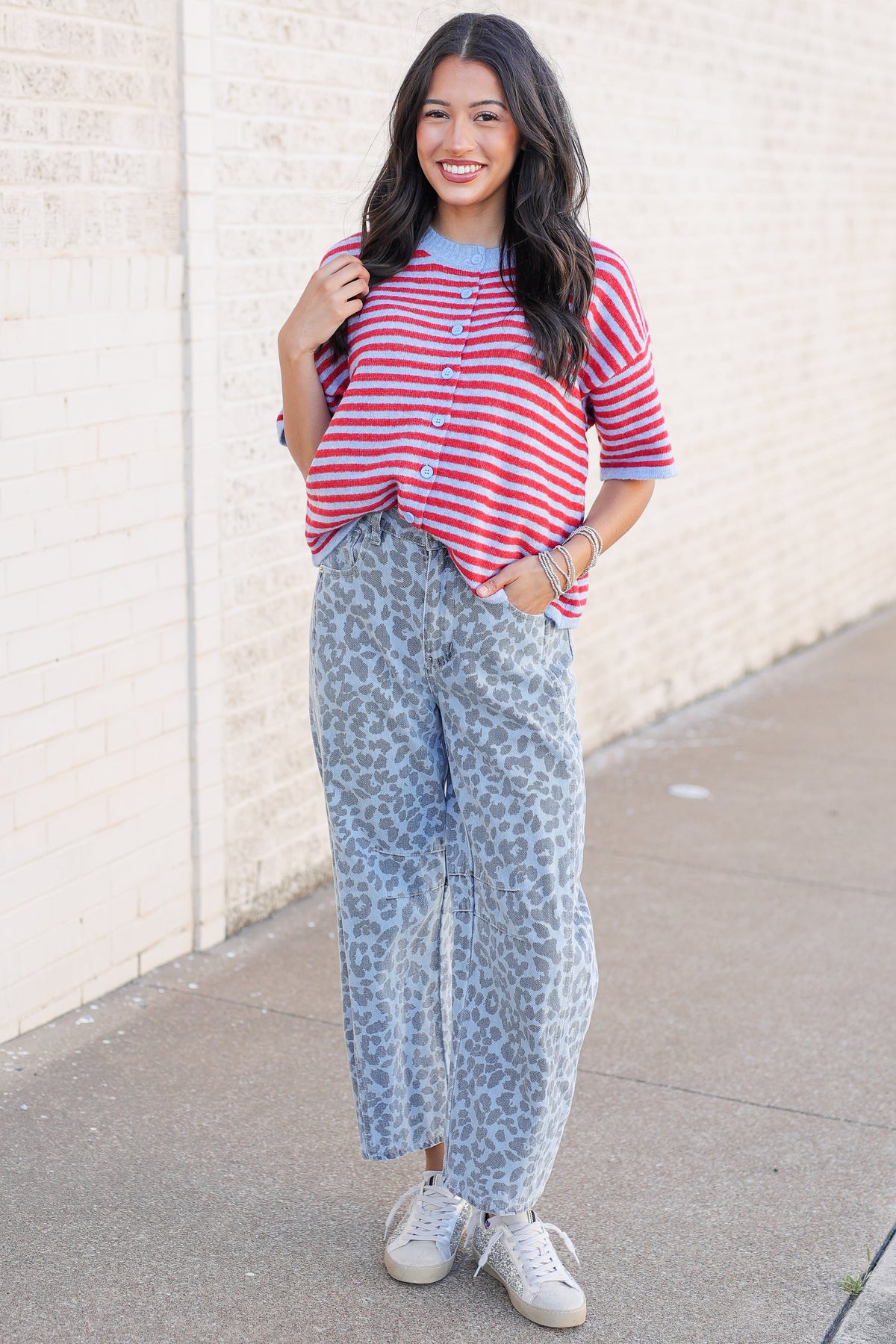 A woman stands outdoors against a light beige brick wall wearing a red and white striped short-sleeve top with blue trim, light blue leopard-print pants, and white sneakers.