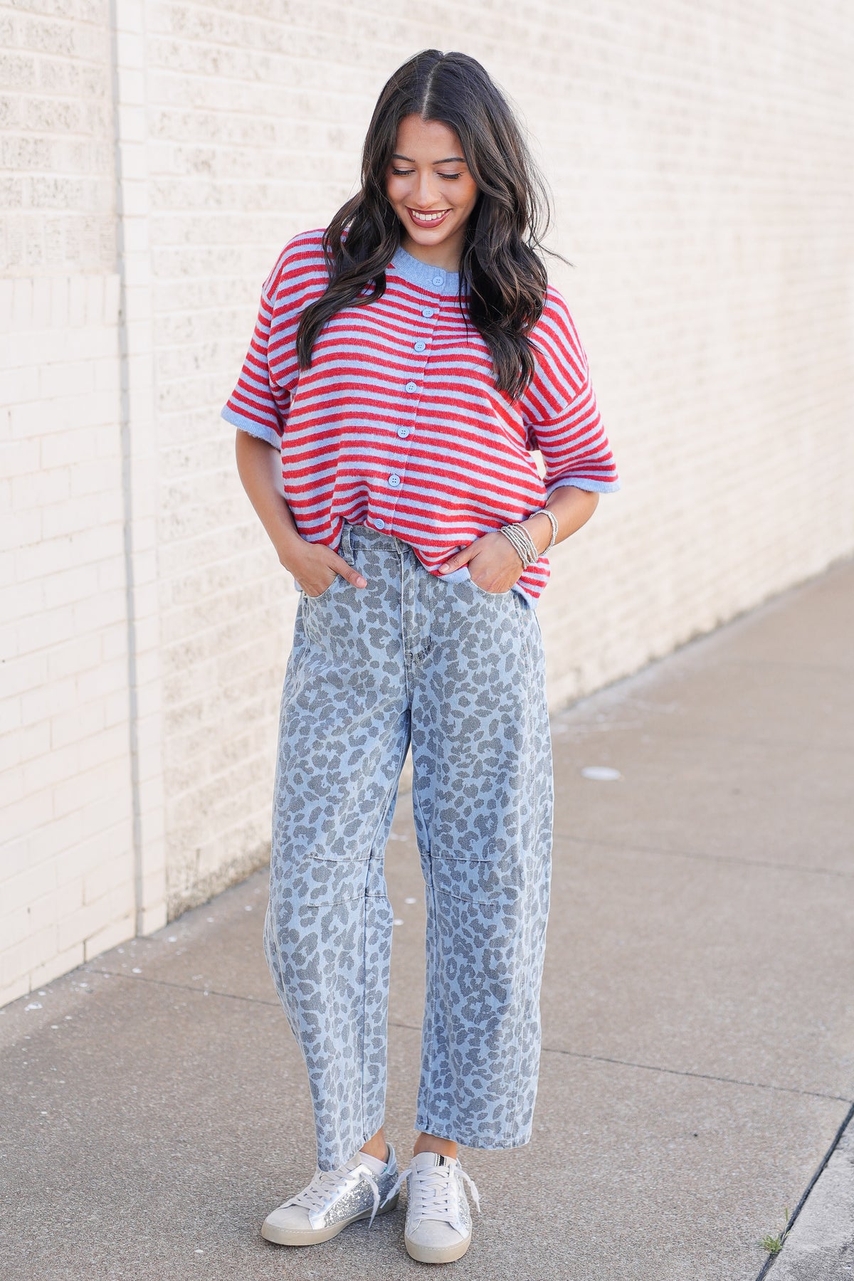 A woman stands outdoors against a light brick wall wearing a red and white striped short-sleeve top, light blue leopard-print wide-leg jeans, and white sneakers.