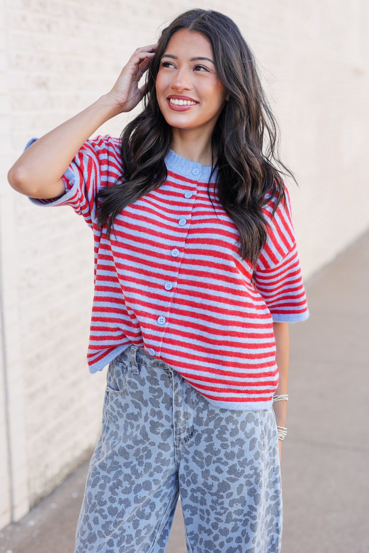 A woman wearing a red and white striped short-sleeve button-up cardigan with a light blue collar and cuffs, paired with gray leopard-print pants, posing outdoors against a beige wall.