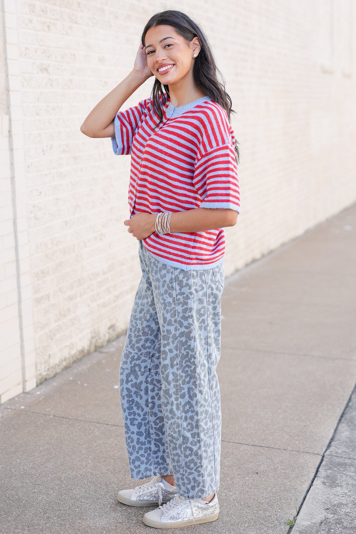 A woman posing outdoors on a sidewalk next to a light-colored brick wall, wearing a red and white striped short-sleeve t-shirt, light blue leopard-print wide-leg pants, and white sneakers.