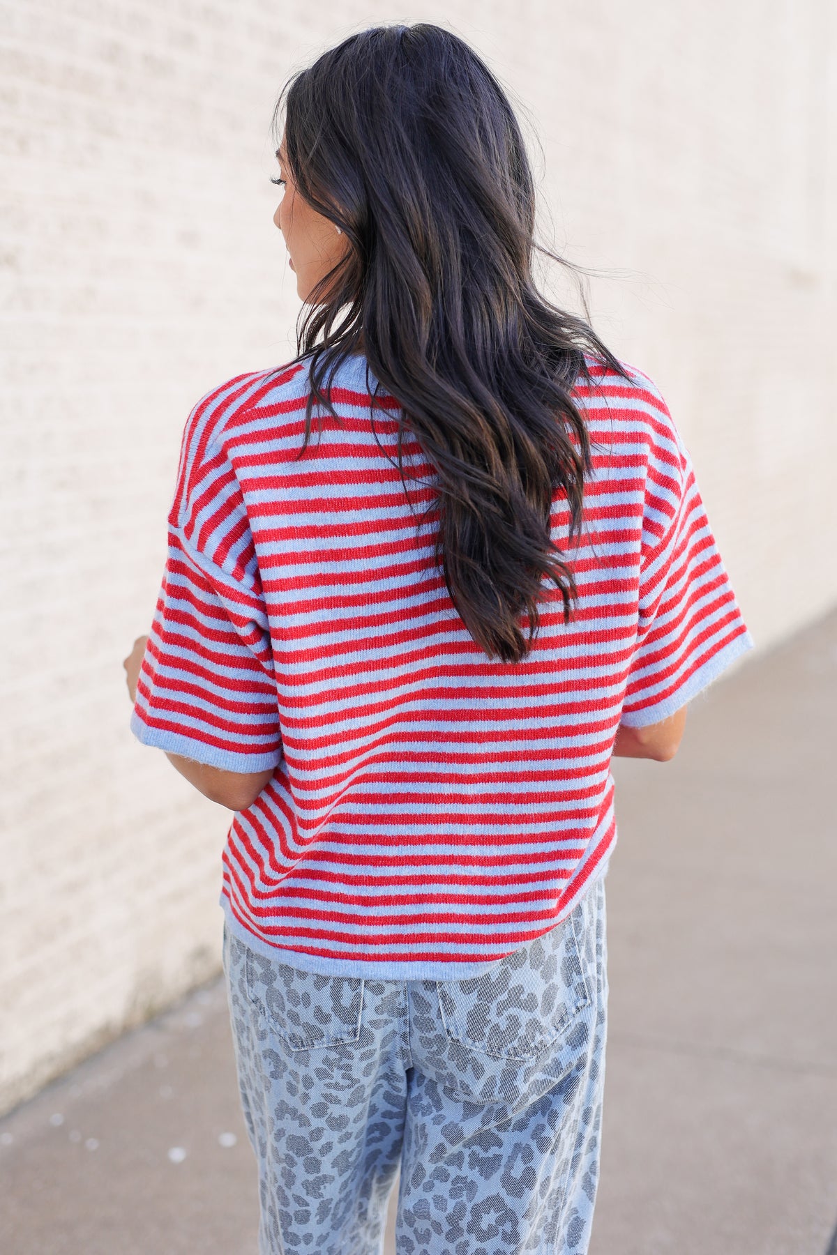 A woman viewed from behind wearing a red and white striped short-sleeve knit top and light blue leopard-print pants, standing outdoors against a light brick wall.