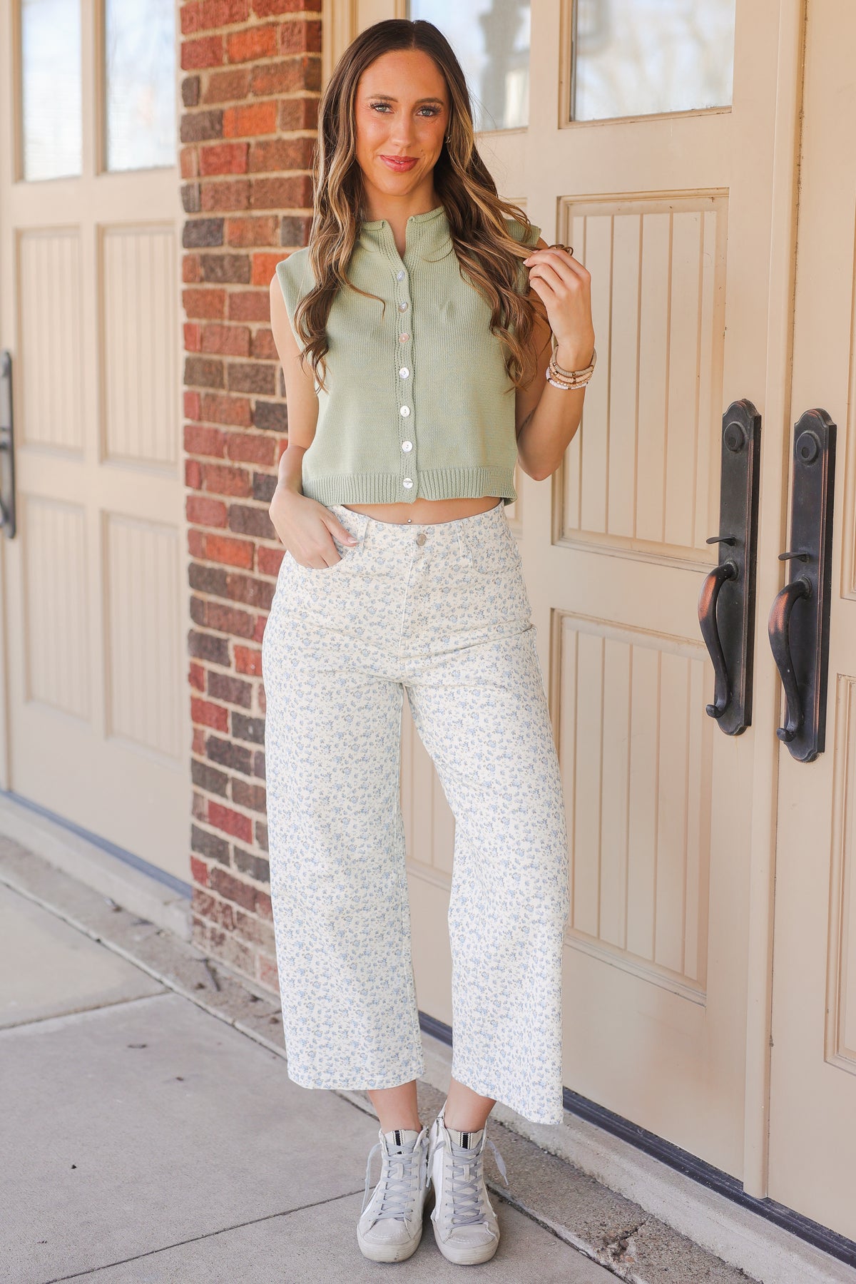 Woman wearing a green top and white patterned pants standing in front of a door.