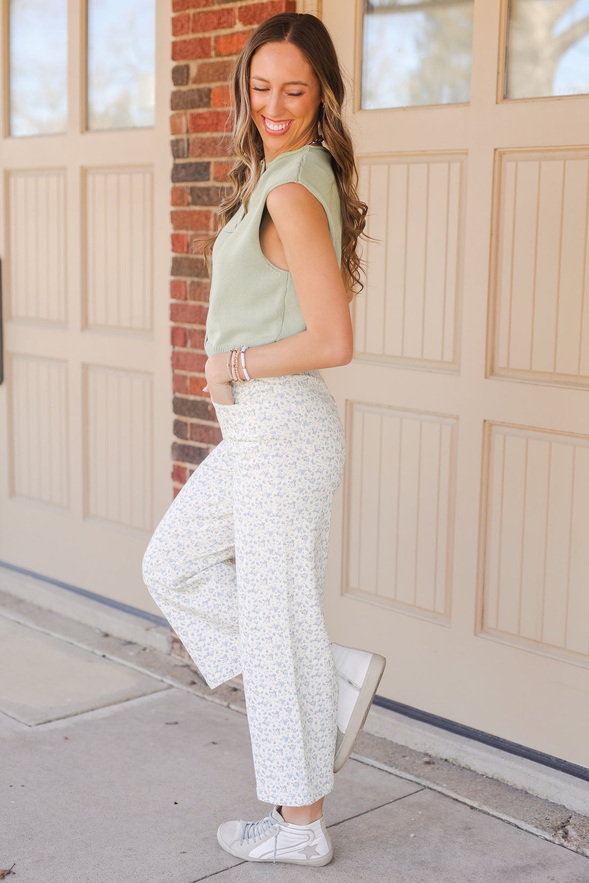 Woman wearing a green sleeveless top and white patterned pants standing in front of a garage door.