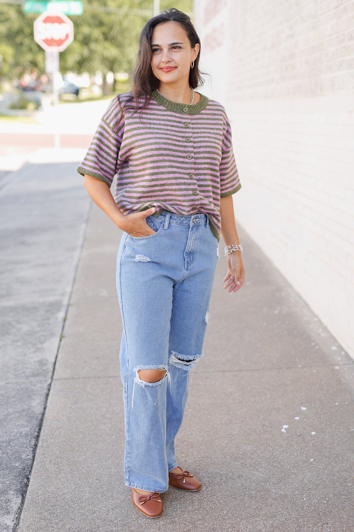A woman stands on a sidewalk wearing a short-sleeve button-front knit top with horizontal stripes in muted red and olive green, paired with light-wash distressed jeans and brown loafers.