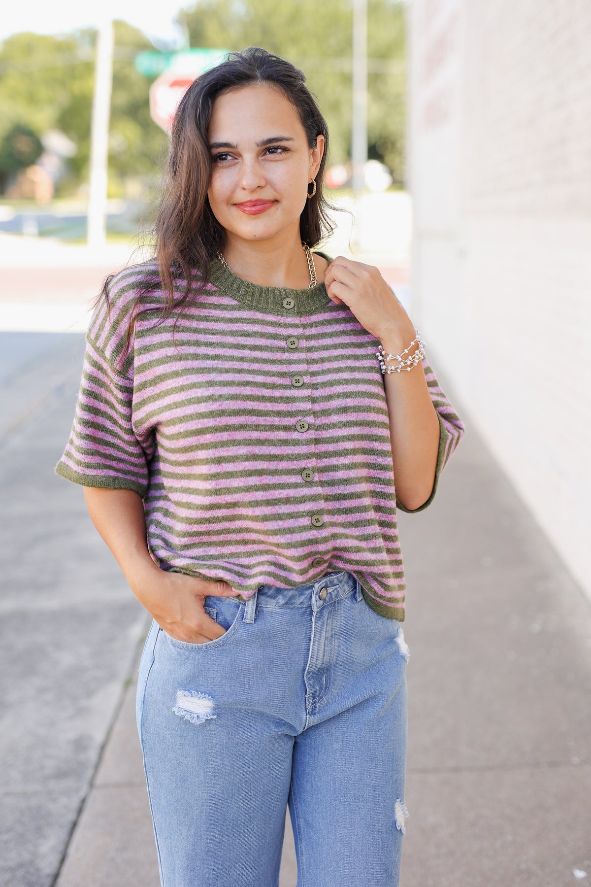 A woman wearing a short-sleeve knit henley top with horizontal olive green and dusty rose stripes, paired with light-wash blue jeans, standing outdoors on a sidewalk.