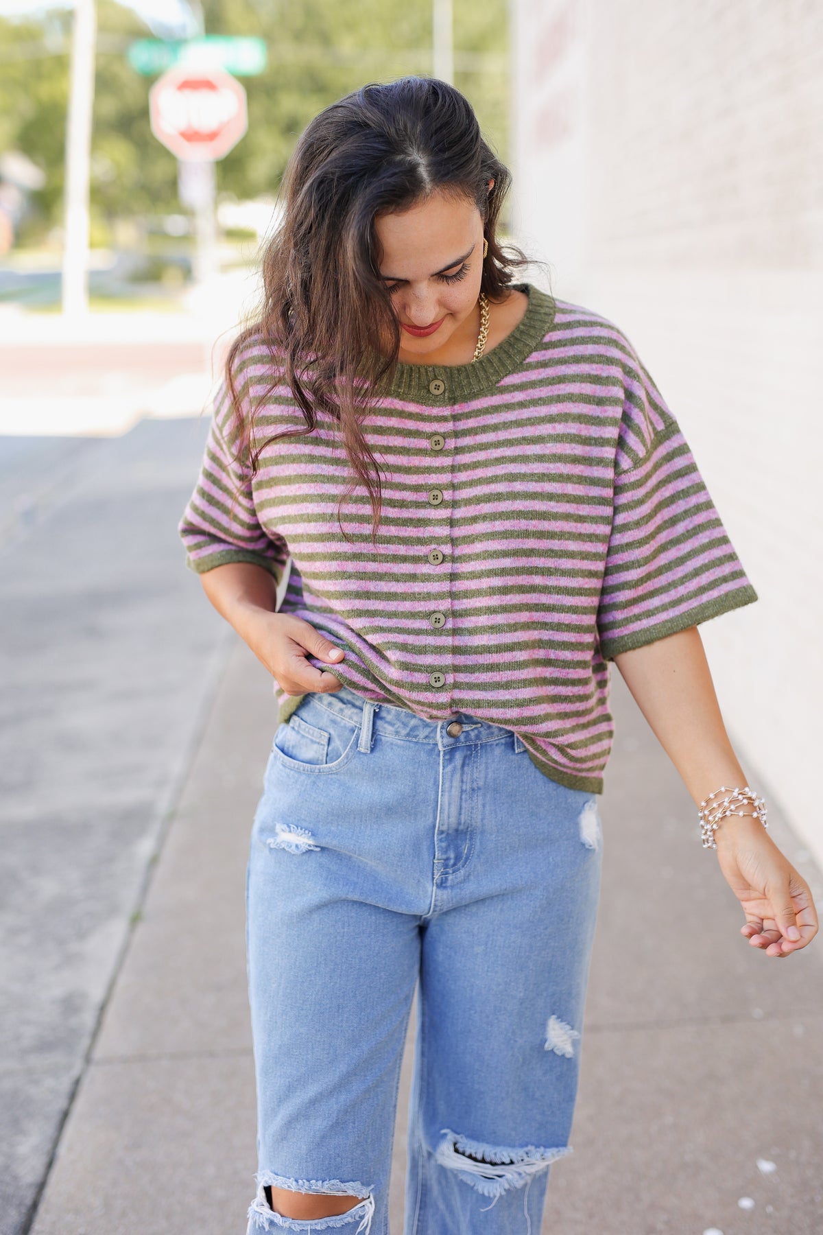A woman wearing a green and brown striped short-sleeve button-up knit top with light-wash distressed jeans, standing on a sidewalk.