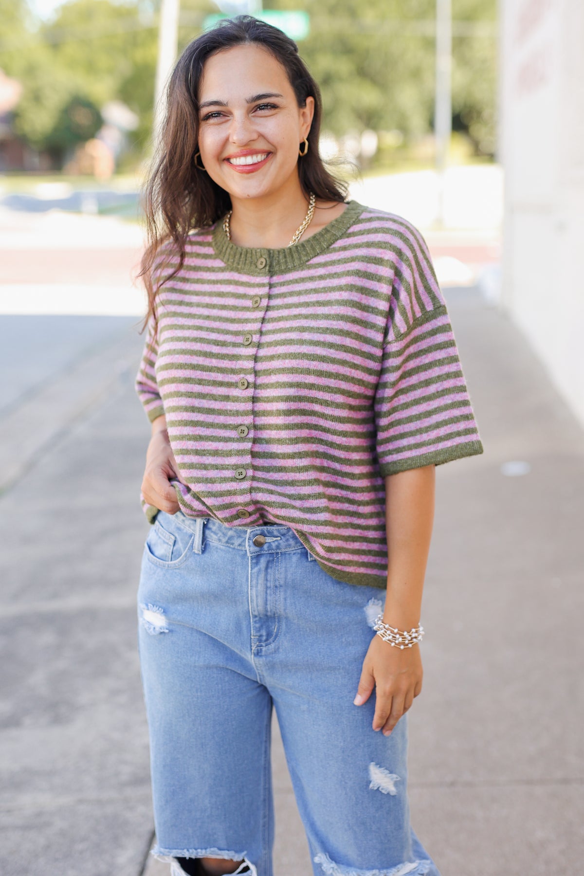 A woman smiling outdoors while wearing a short-sleeve knit top with horizontal olive green and dusty rose stripes, paired with light blue distressed jeans.
