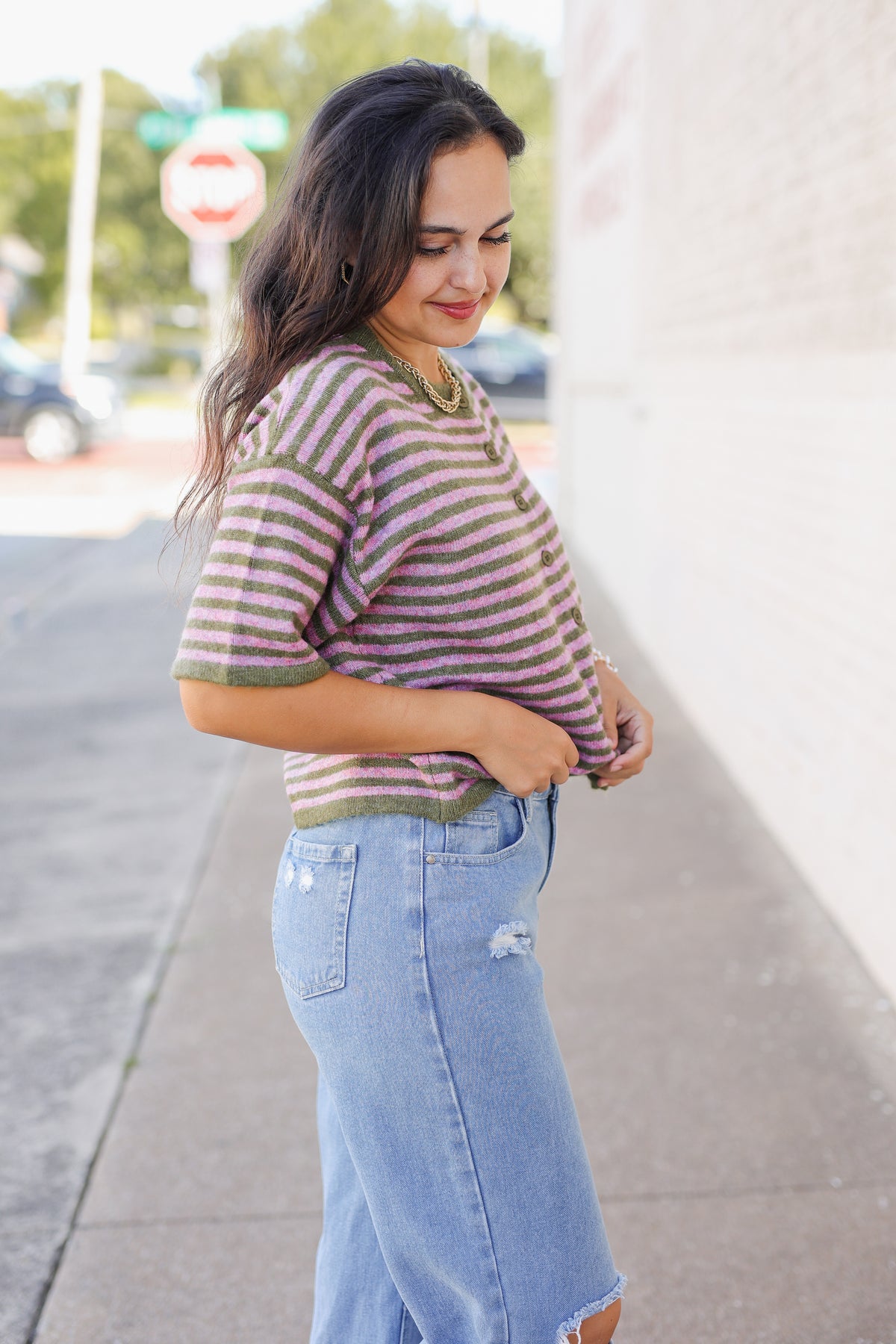 A woman wearing a short-sleeve striped knit top with olive green and dusty rose stripes, paired with light-wash distressed denim jeans, standing on a sidewalk next to a white brick wall.