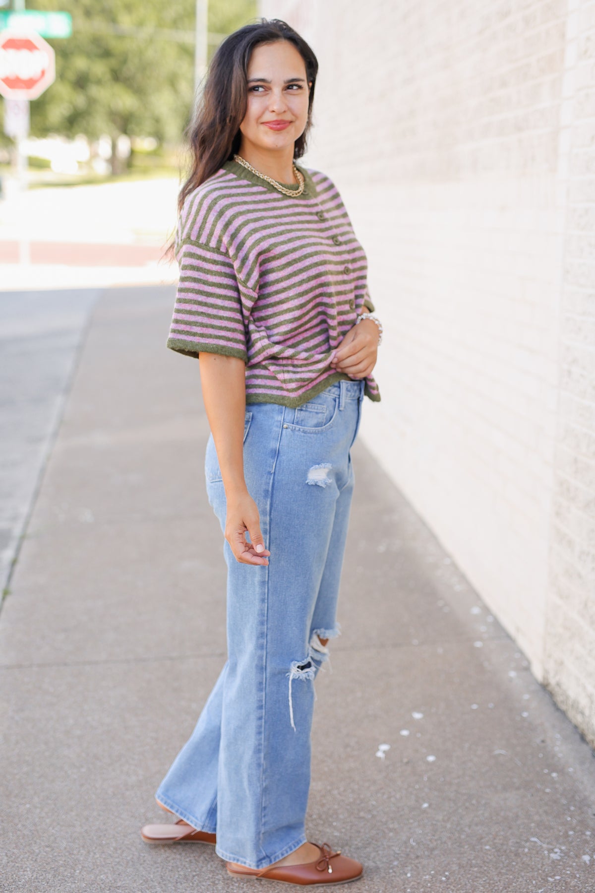 A woman stands on a sidewalk wearing a short-sleeve striped knit top with olive green trim, light blue distressed flare jeans, and brown flat shoes, posed against a light brick wall.