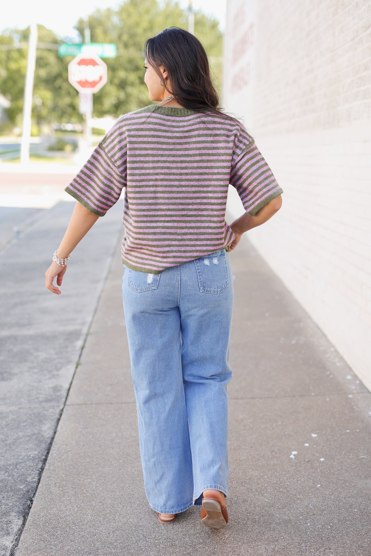 A woman is shown from behind wearing a short-sleeve knit top with horizontal brown and beige stripes, olive green trim, and light blue wide-leg jeans, walking on a sidewalk.
