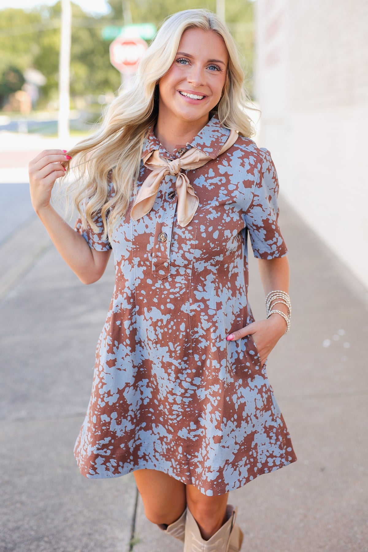 A woman wearing a short-sleeve brown and light blue tie-dye dress with a beige necktie, standing outdoors on a paved walkway.