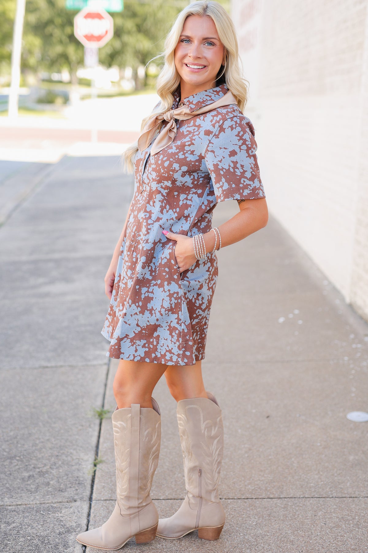 A woman modeling a short-sleeve brown and light blue floral print dress with a tan neck scarf, paired with beige cowboy boots, standing on a sidewalk.