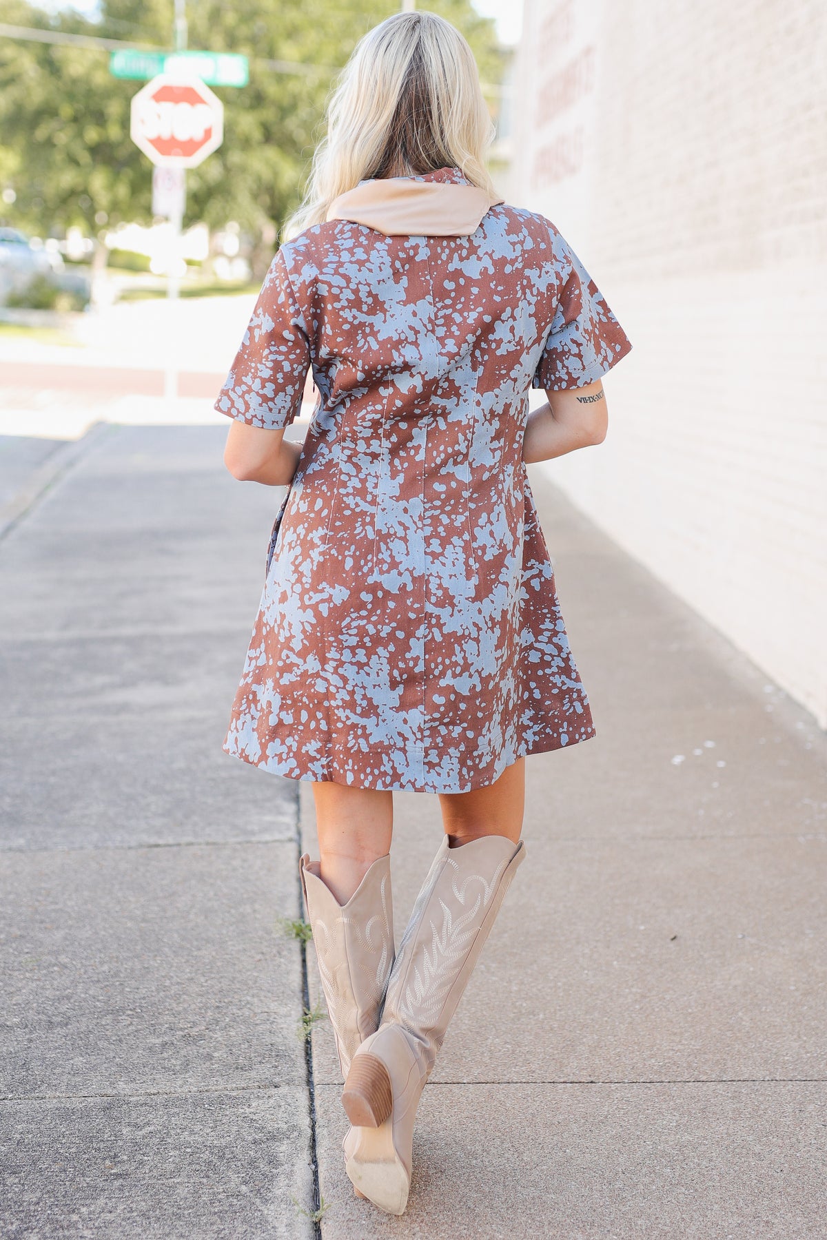A woman viewed from behind wearing a brown and light blue floral print short-sleeve dress with a beige collar, paired with beige cowboy boots, walking on a sidewalk.