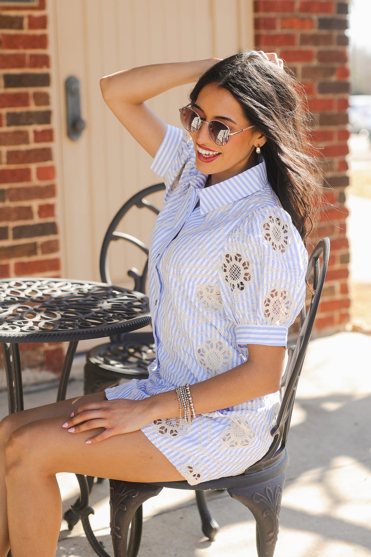 Woman sitting outdoors wearing a light blue top and bottom with lace details, sunglasses, and a bracelet.