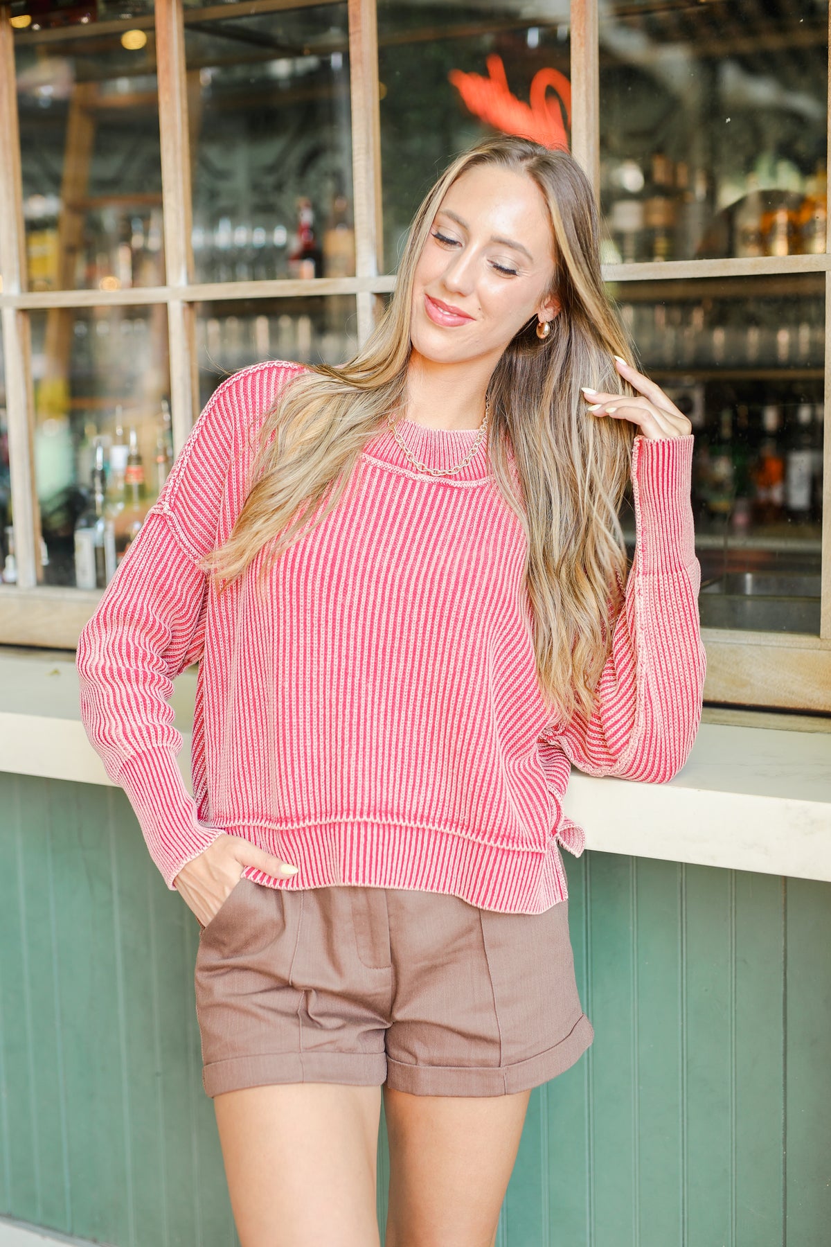 Woman wearing a pink striped sweater and brown shorts standing in front of a bar.