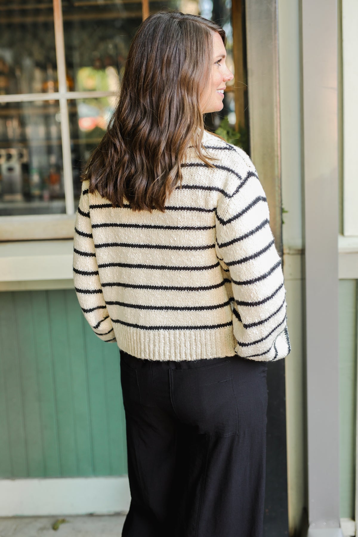 Woman wearing a striped sweater and black pants standing in front of a window.