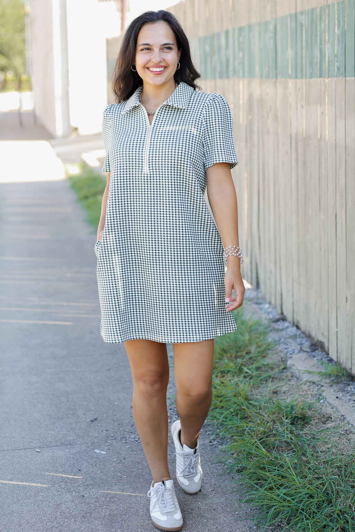 A woman wearing a short-sleeve gingham mini dress with a half-zip collar and side pockets, walking outdoors on a paved path next to a wooden fence.