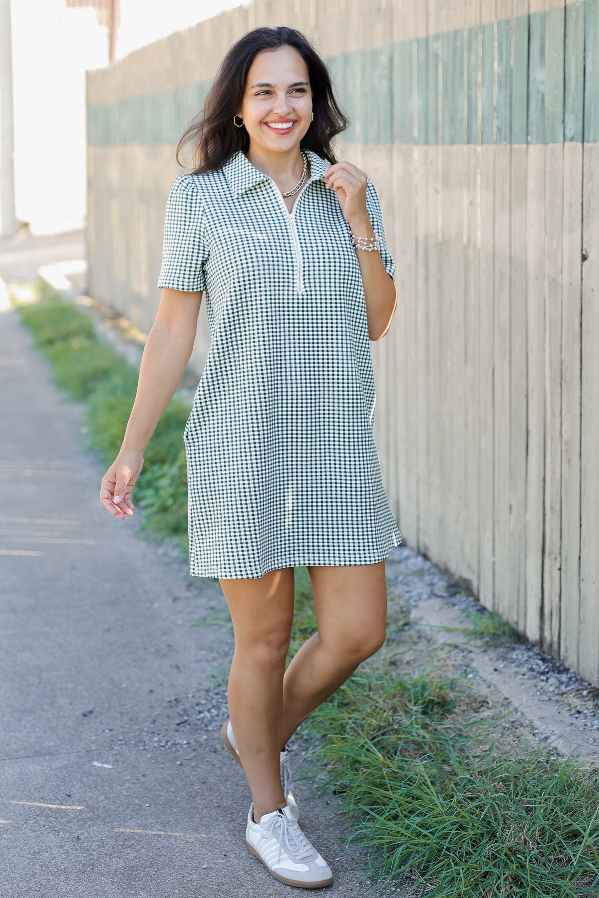 A woman wearing a short-sleeve gingham mini dress with a half-zip collar and side pockets, walking outdoors on a paved path