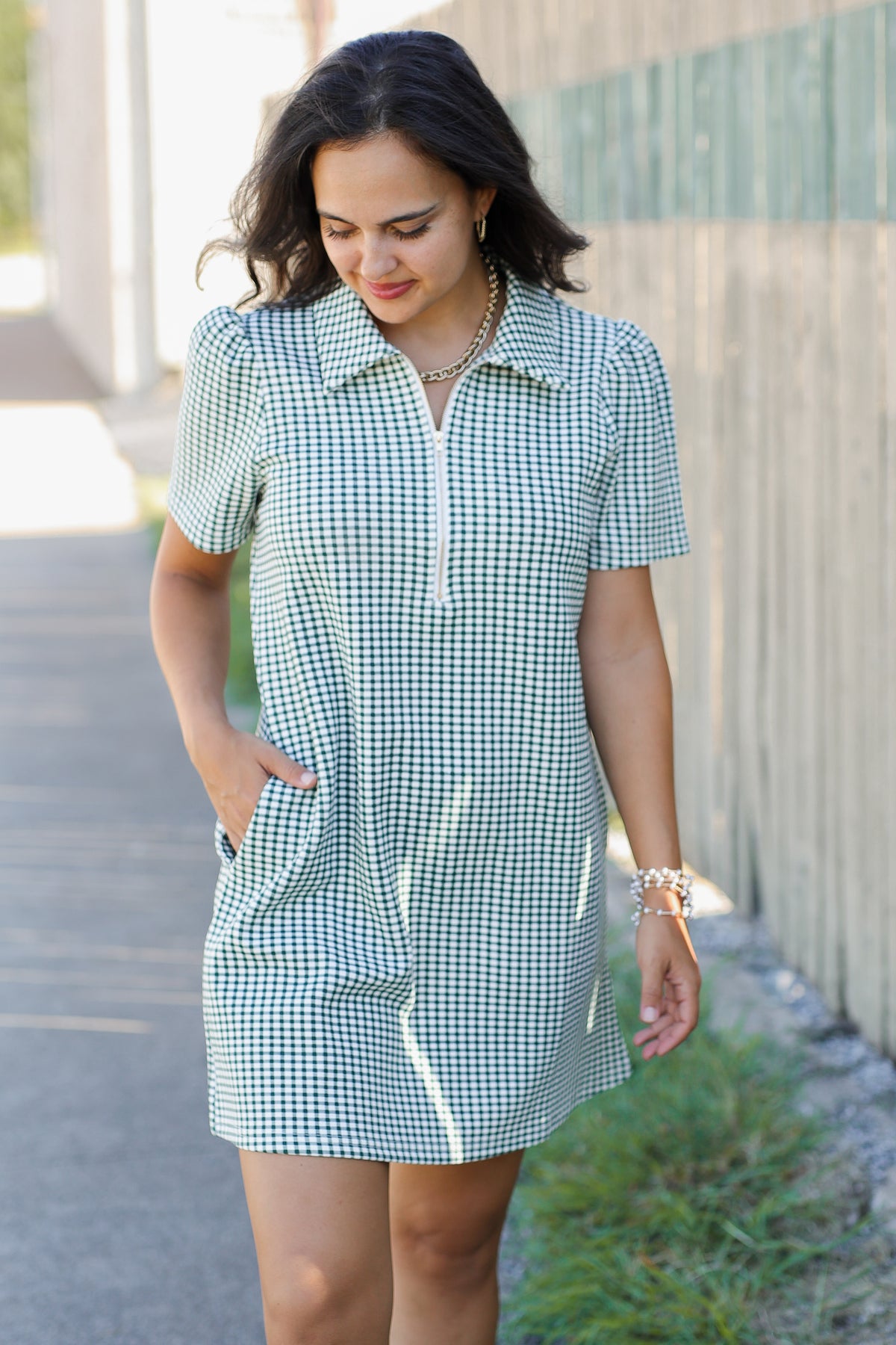 A woman wearing a short-sleeve green and white gingham mini dress with a collar and front pockets, standing outdoors against a concrete wall.