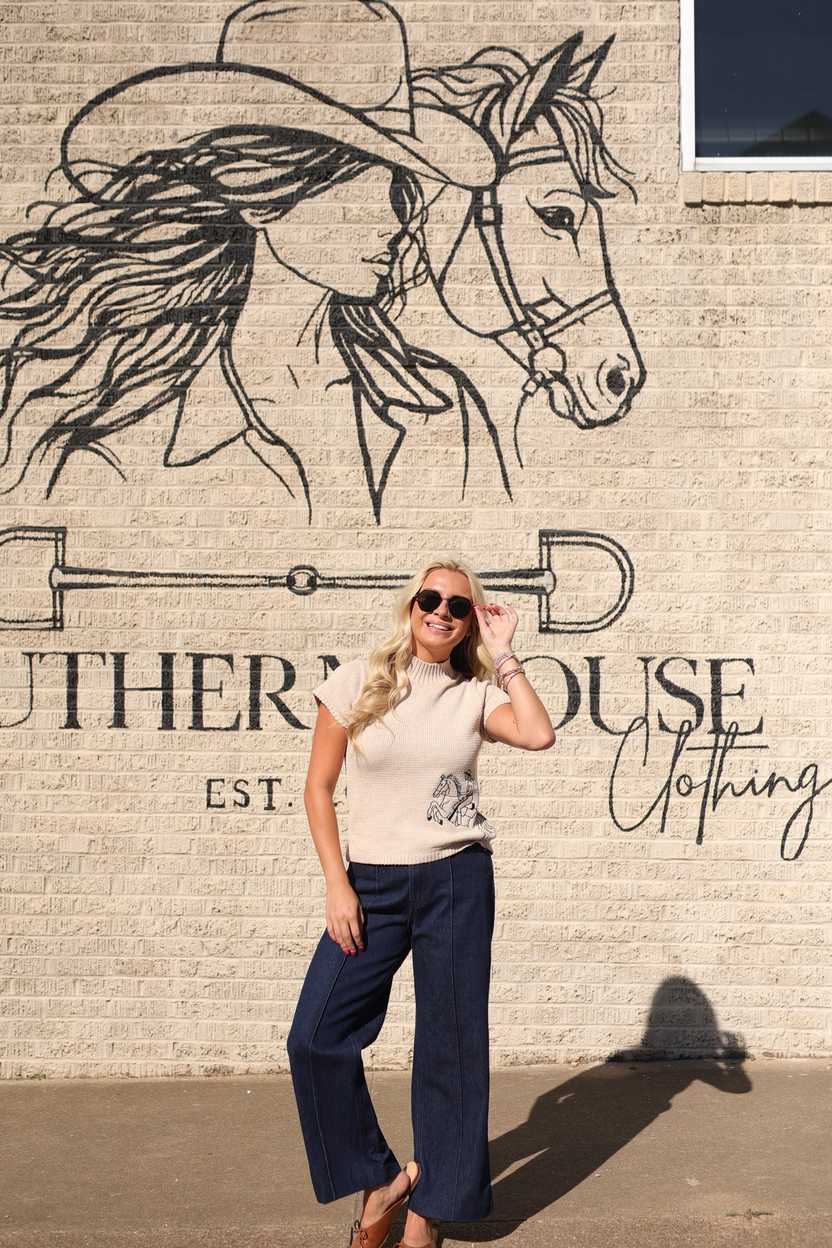 A woman posing in front of a brick wall with a large black line-art mural of a horse wearing a cowboy hat and the text 'Southern House Clothing'. She is wearing a cream short-sleeve knit top with a small graphic print on the front and dark navy wide-leg pants.
