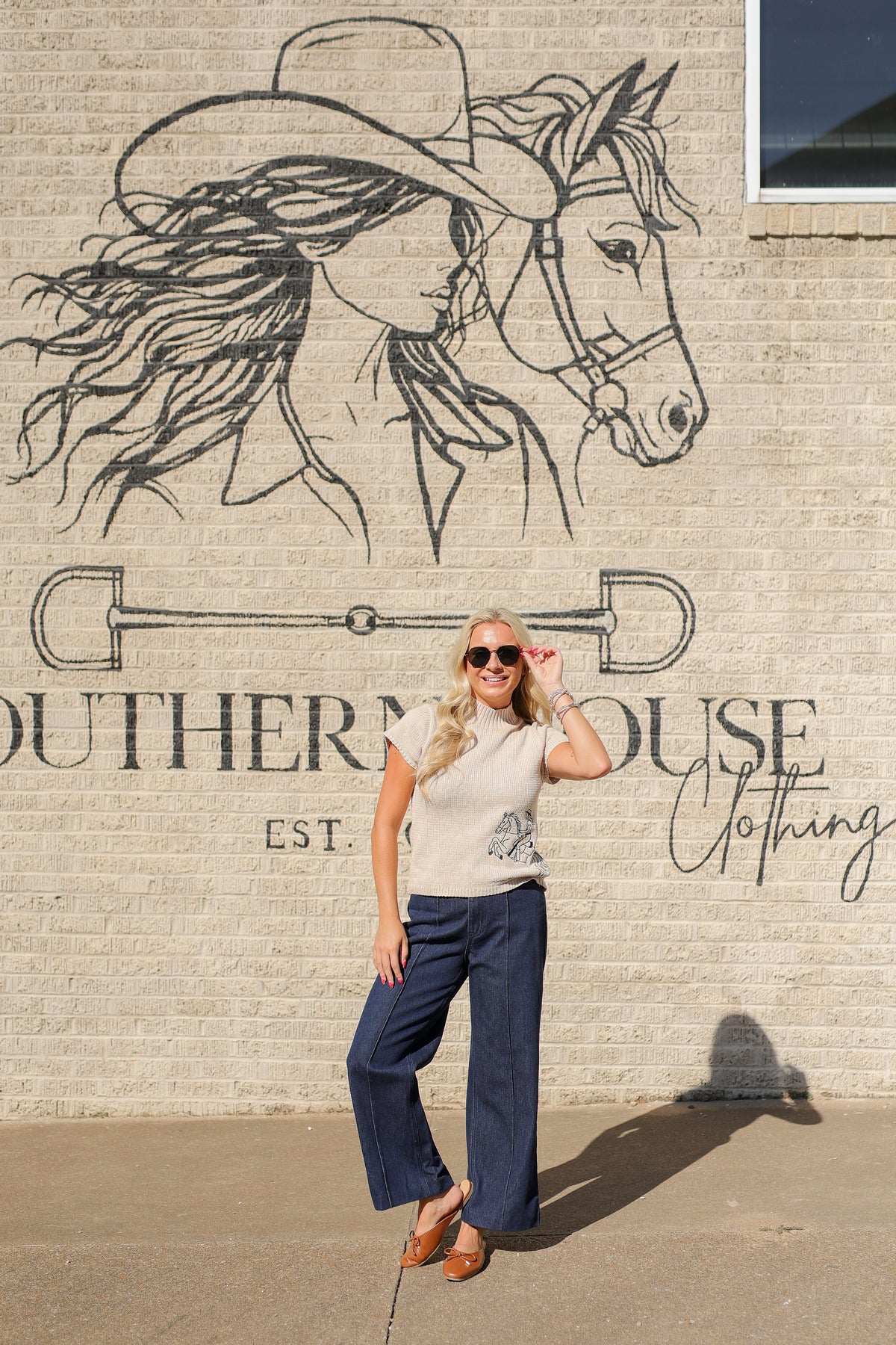 A woman posing in front of a brick wall with a large western-themed mural. She is wearing a cream short-sleeve t-shirt, dark blue wide-leg pants, and brown leather flat shoes.