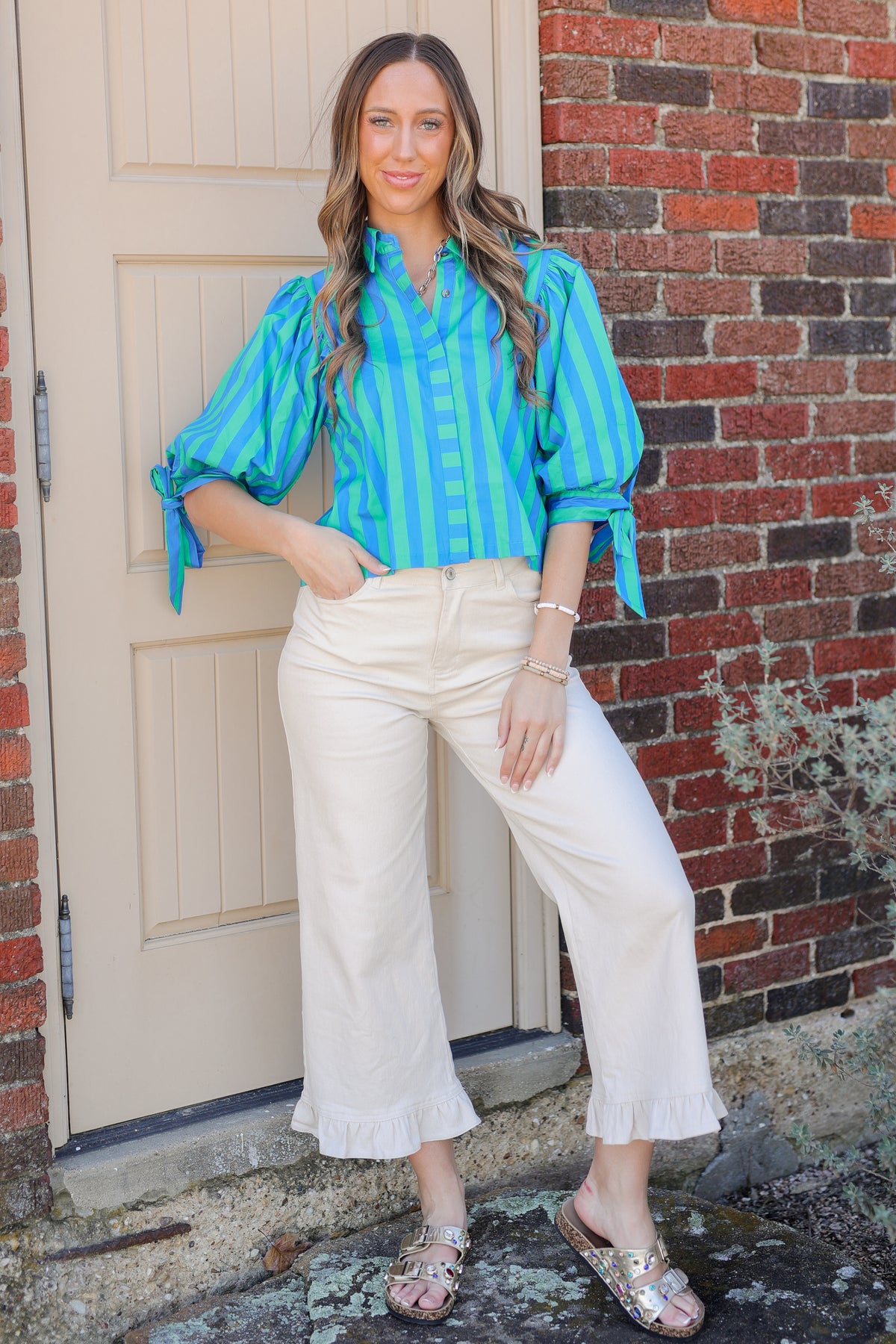 Woman wearing a blue striped shirt and white pants standing in front of a brick wall.