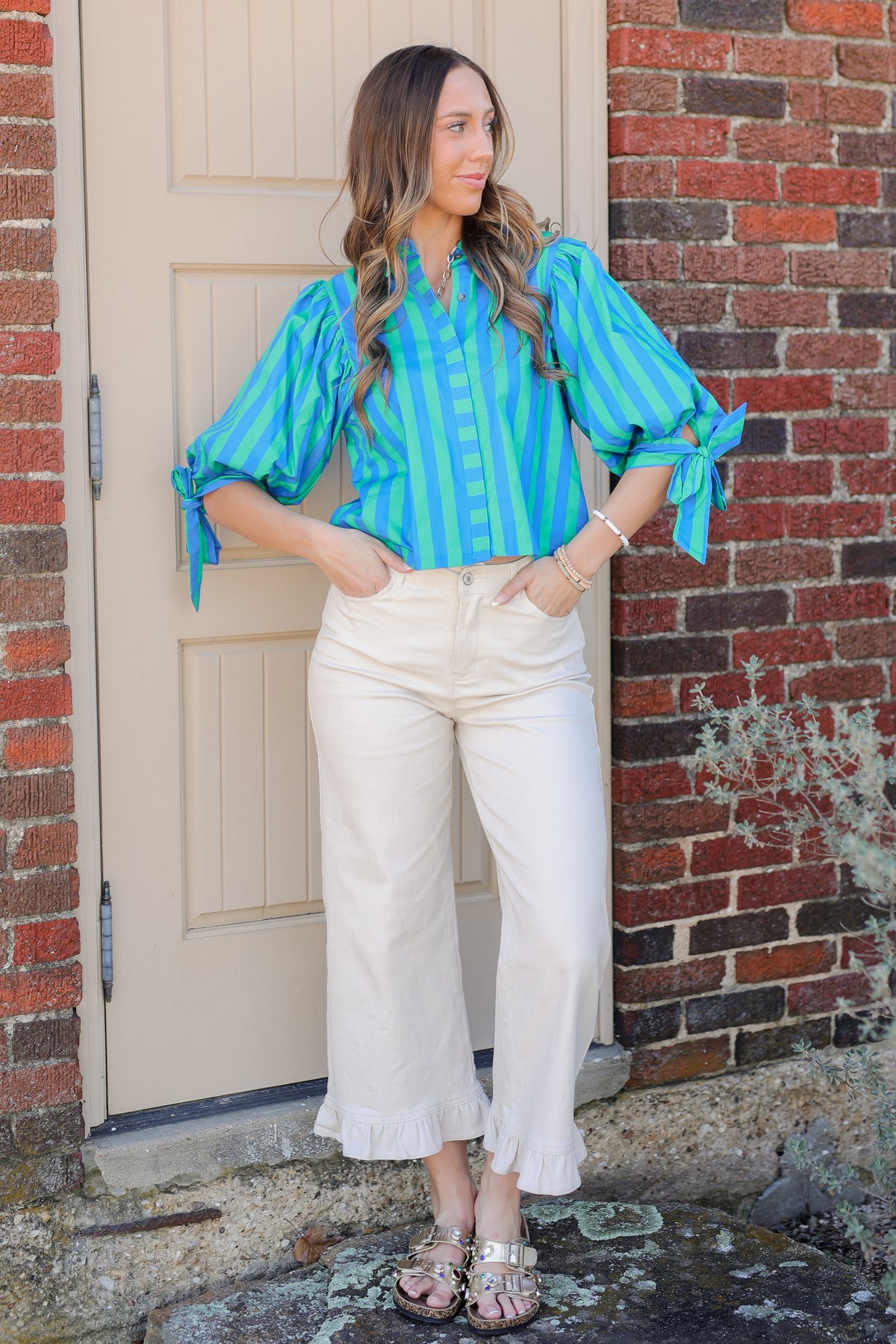 Woman wearing a blue striped blouse and white pants standing in front of a brick wall.