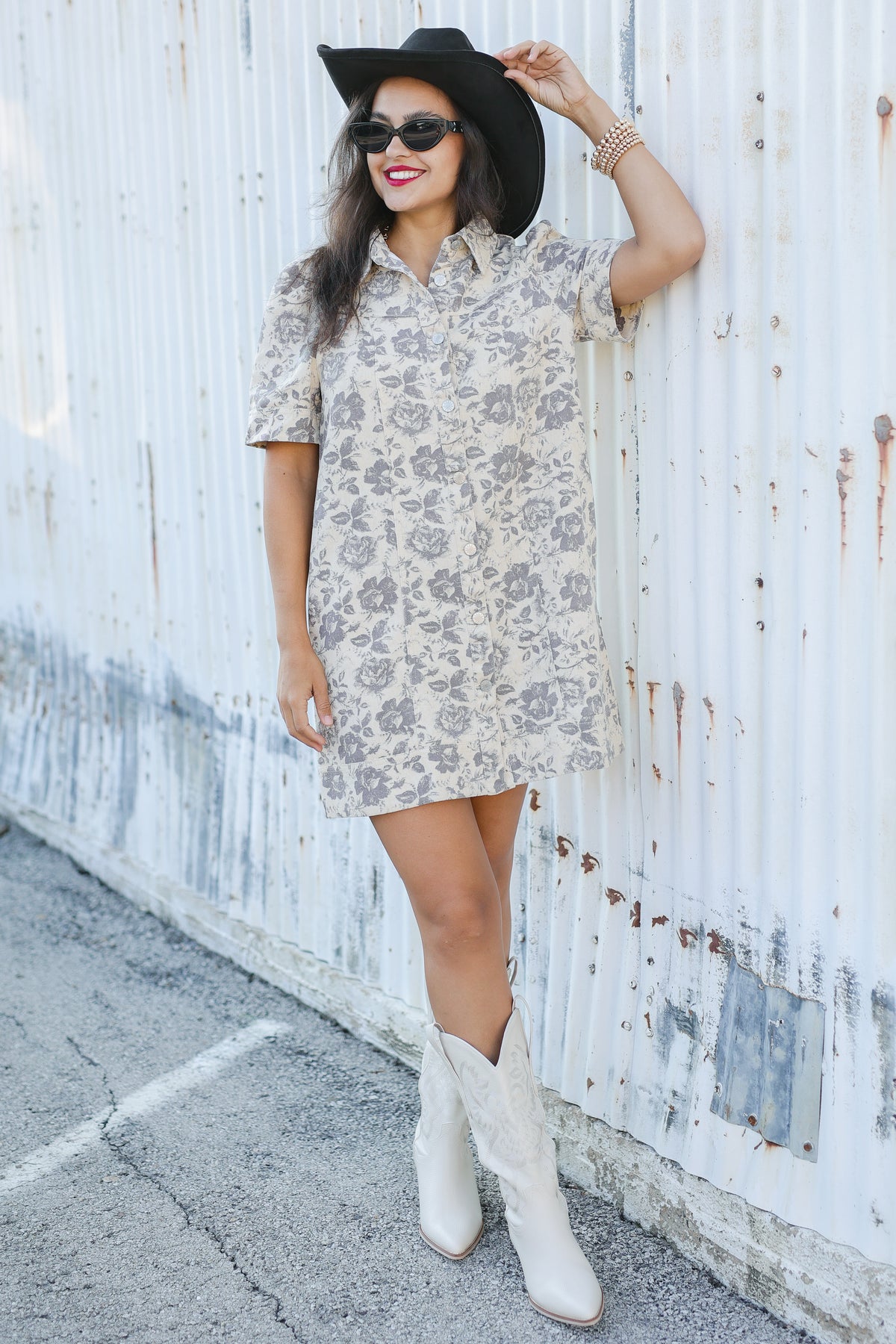 Woman wearing a floral dress, black hat, and white boots against a white wall.