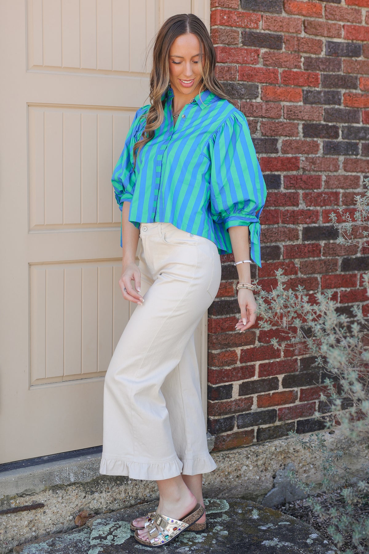 Woman wearing a blue and green striped shirt and white pants standing against a brick wall.