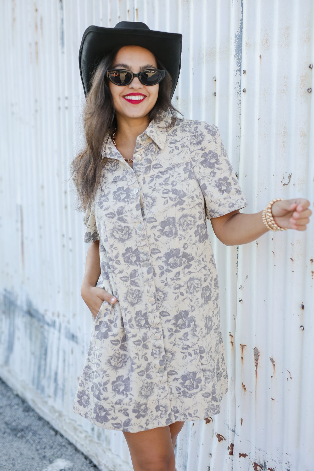 Woman wearing a floral dress, black hat, and sunglasses against a white wall.