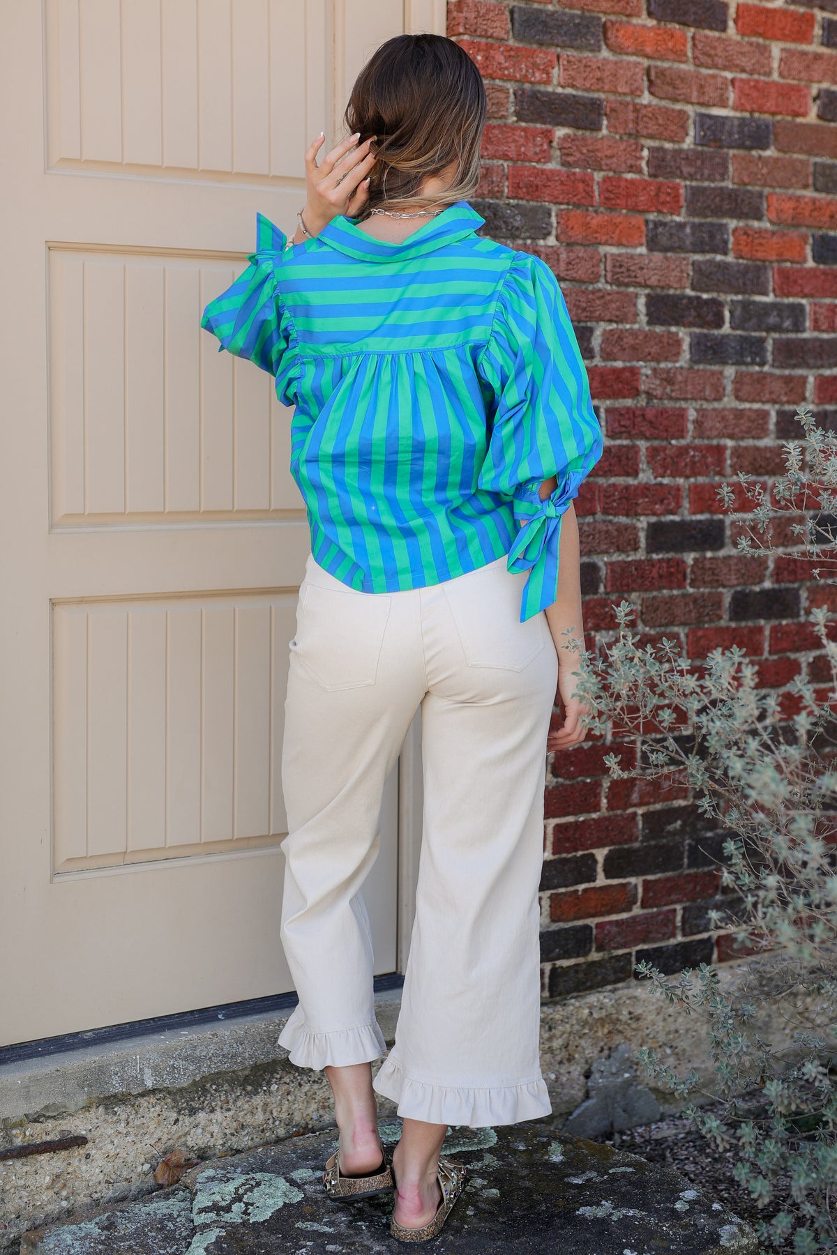 Person wearing a blue and green striped shirt and white pants standing against a brick wall.