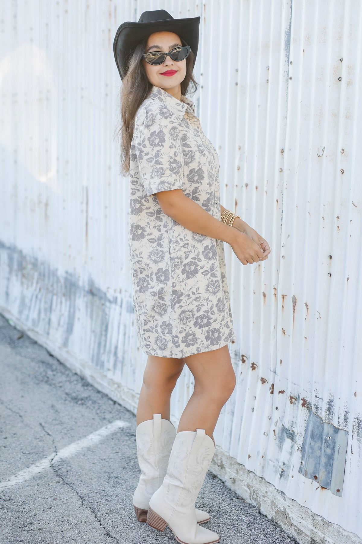 Woman in a floral dress and white cowboy boots standing against a white wall.