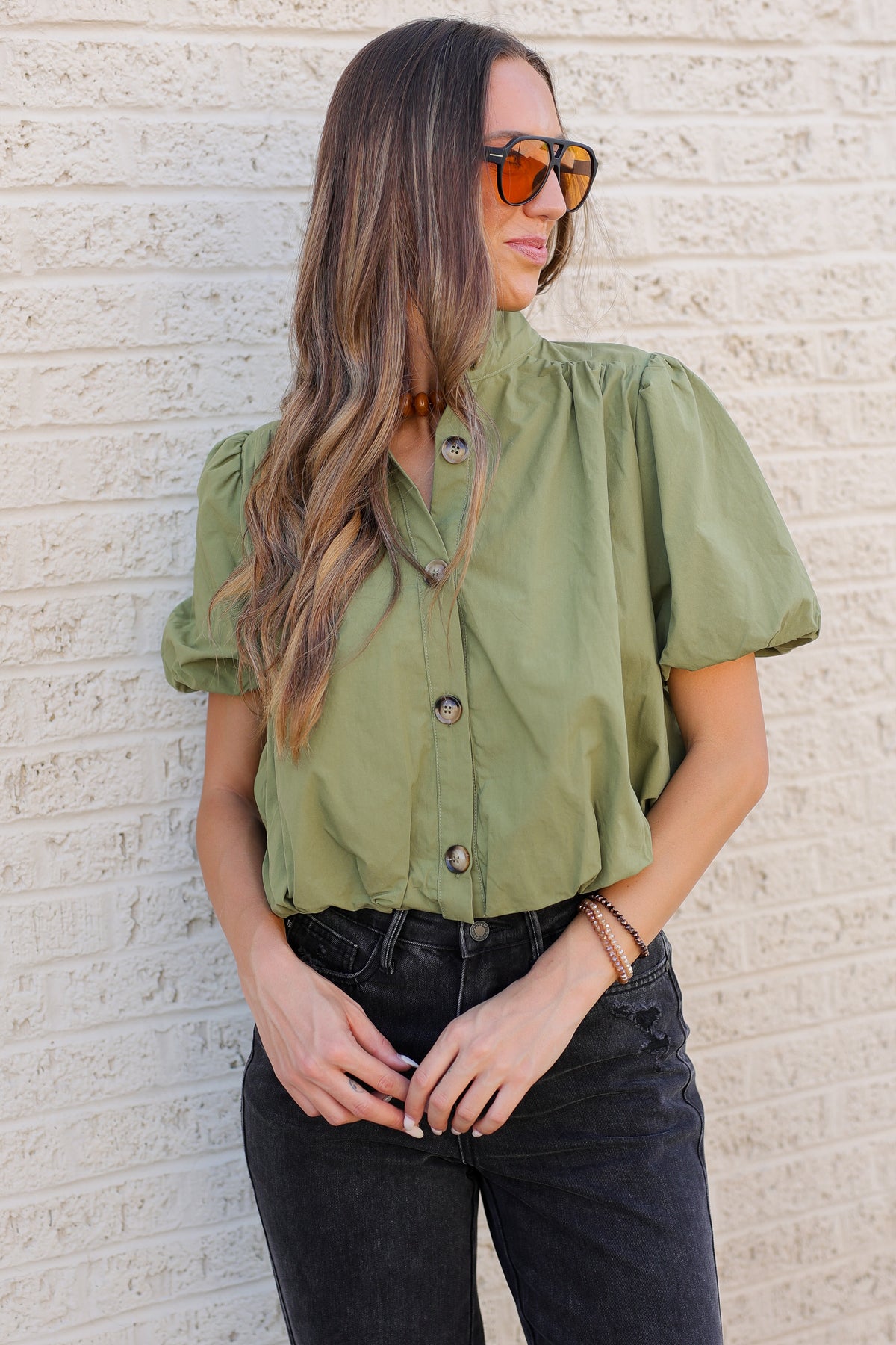 A woman stands against a light beige brick wall wearing an olive green short-sleeve button-up shirt, black distressed jeans, white loafers, and orange-tinted sunglasses.