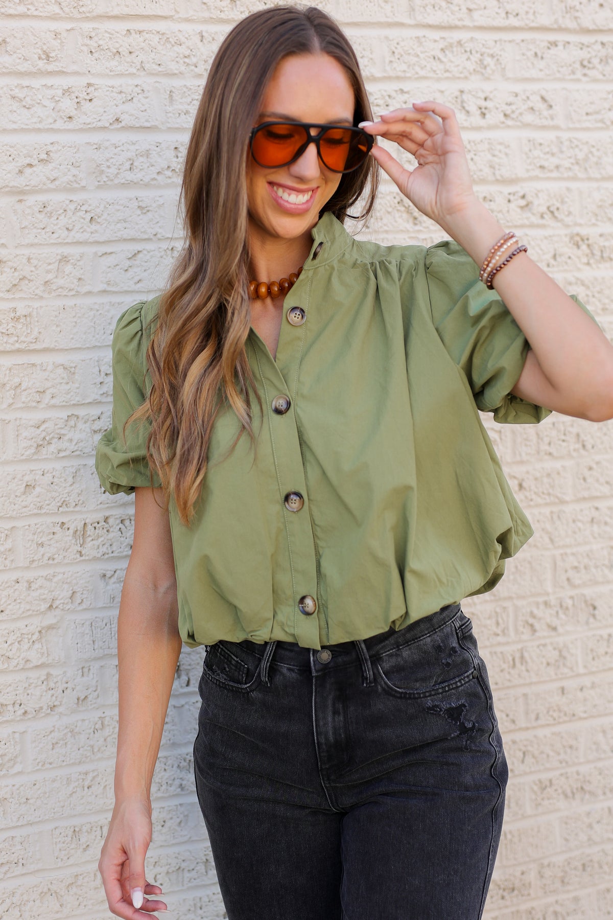 A woman wearing an olive green short-sleeve button-up blouse with puff sleeves, paired with black jeans and tortoiseshell sunglasses, standing against a light beige textured brick wall.