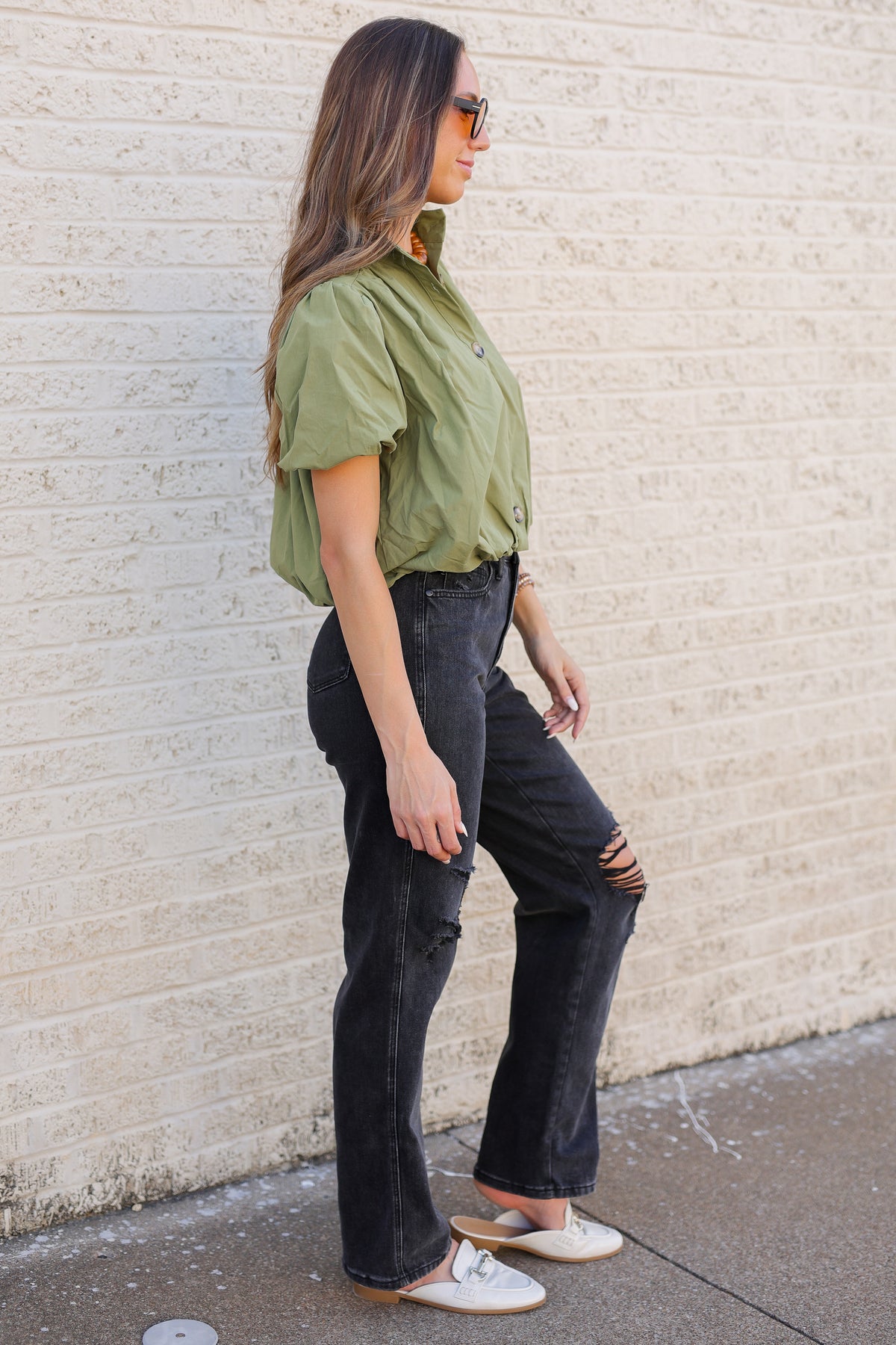 A woman stands in profile against a light beige brick wall, wearing an olive green short-sleeve button-up shirt tucked into black wide-leg distressed jeans with white slip-on shoes.