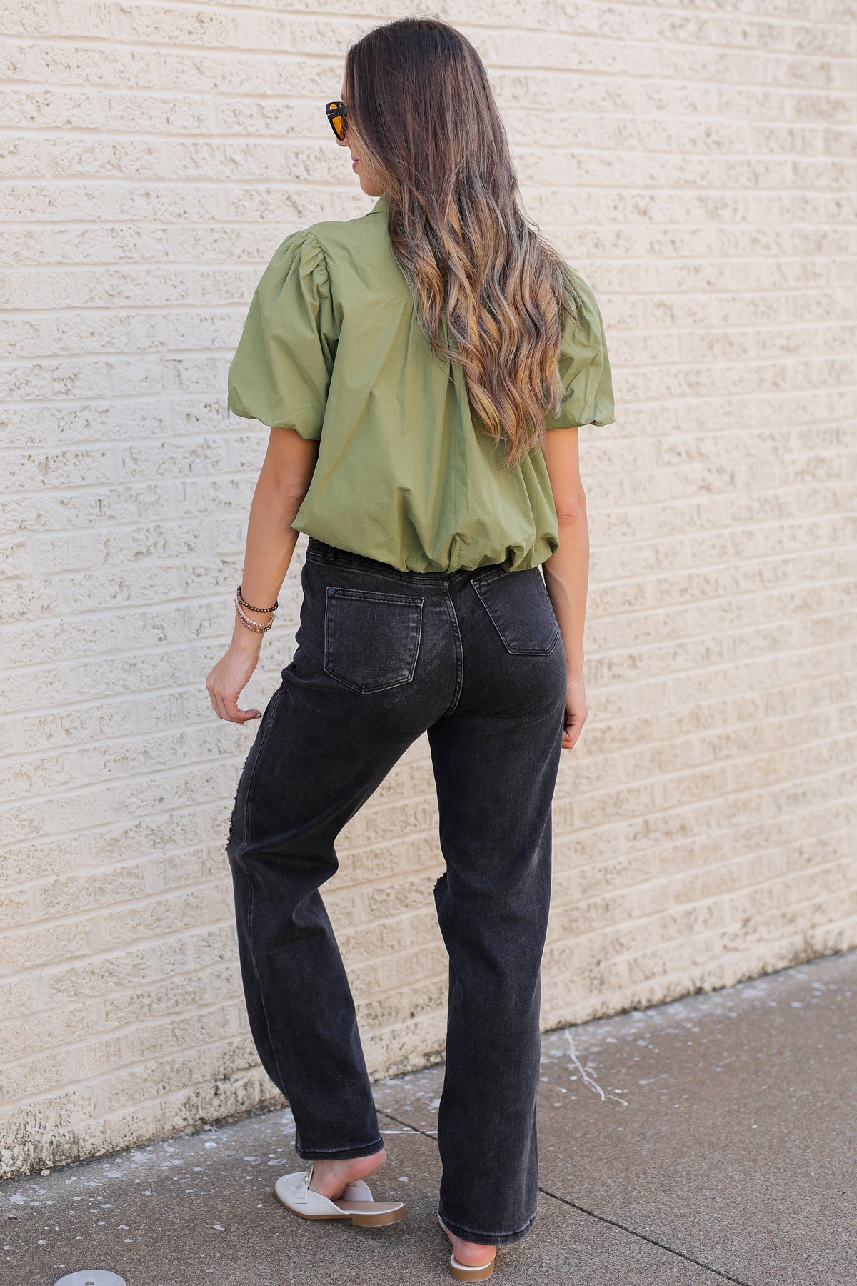 A woman is shown from behind wearing an olive green puff-sleeve blouse and black wide-leg jeans, standing on a sidewalk against a light beige brick wall.