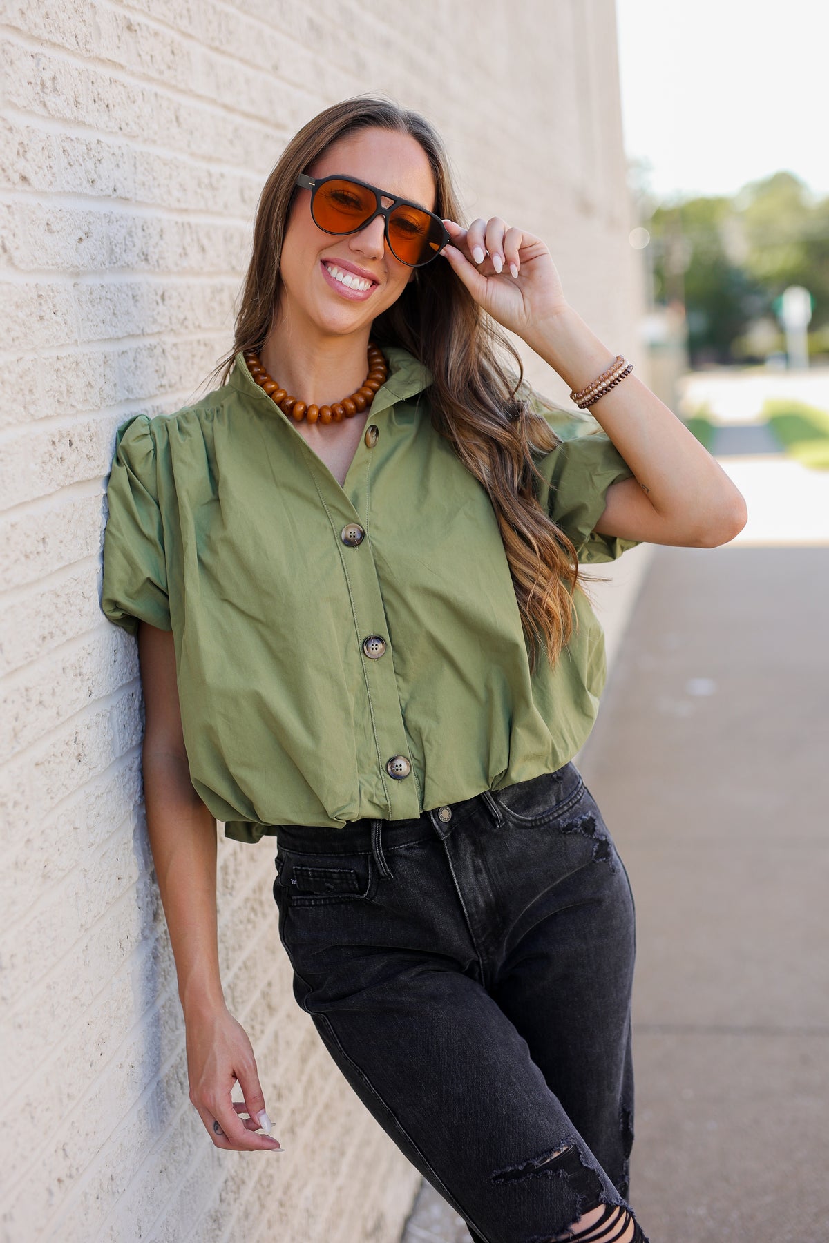 A woman wearing an olive green short-sleeve button-up shirt and black jeans leans against a light brick wall outdoors. She is wearing orange-tinted sunglasses and a chunky brown beaded necklace.