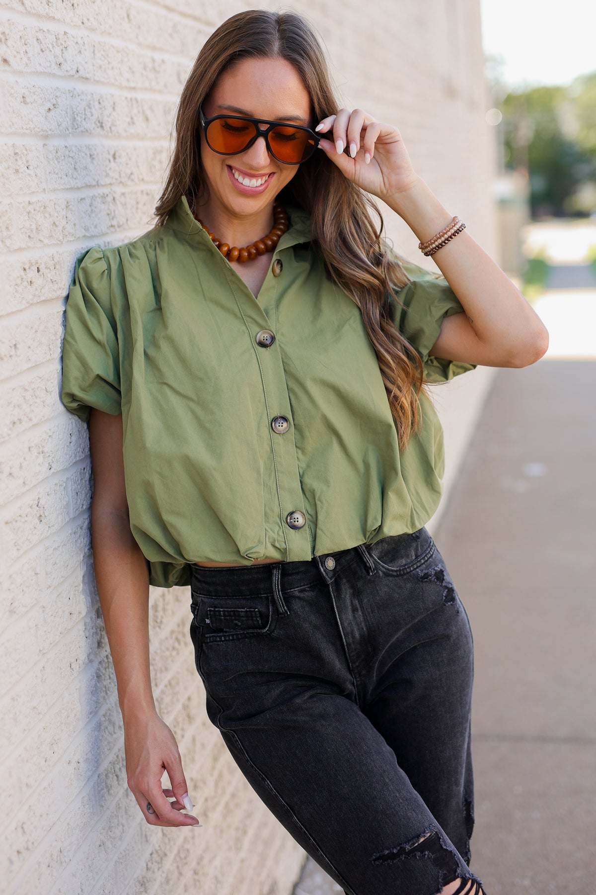 A woman wearing an olive green short-sleeve button-up shirt and black jeans, leaning against a light brick wall outdoors.