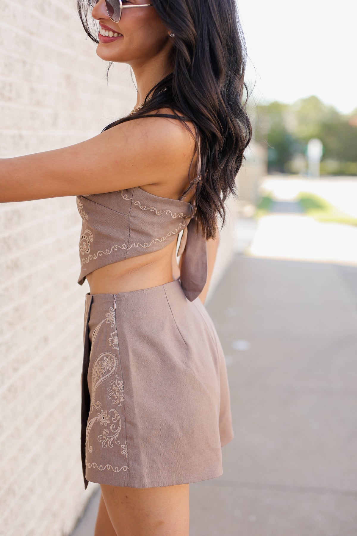A woman wearing a matching taupe two-piece outfit consisting of a sleeveless crop top with lace trim and matching high-waisted shorts with embroidery, standing outdoors next to a light brick wall.