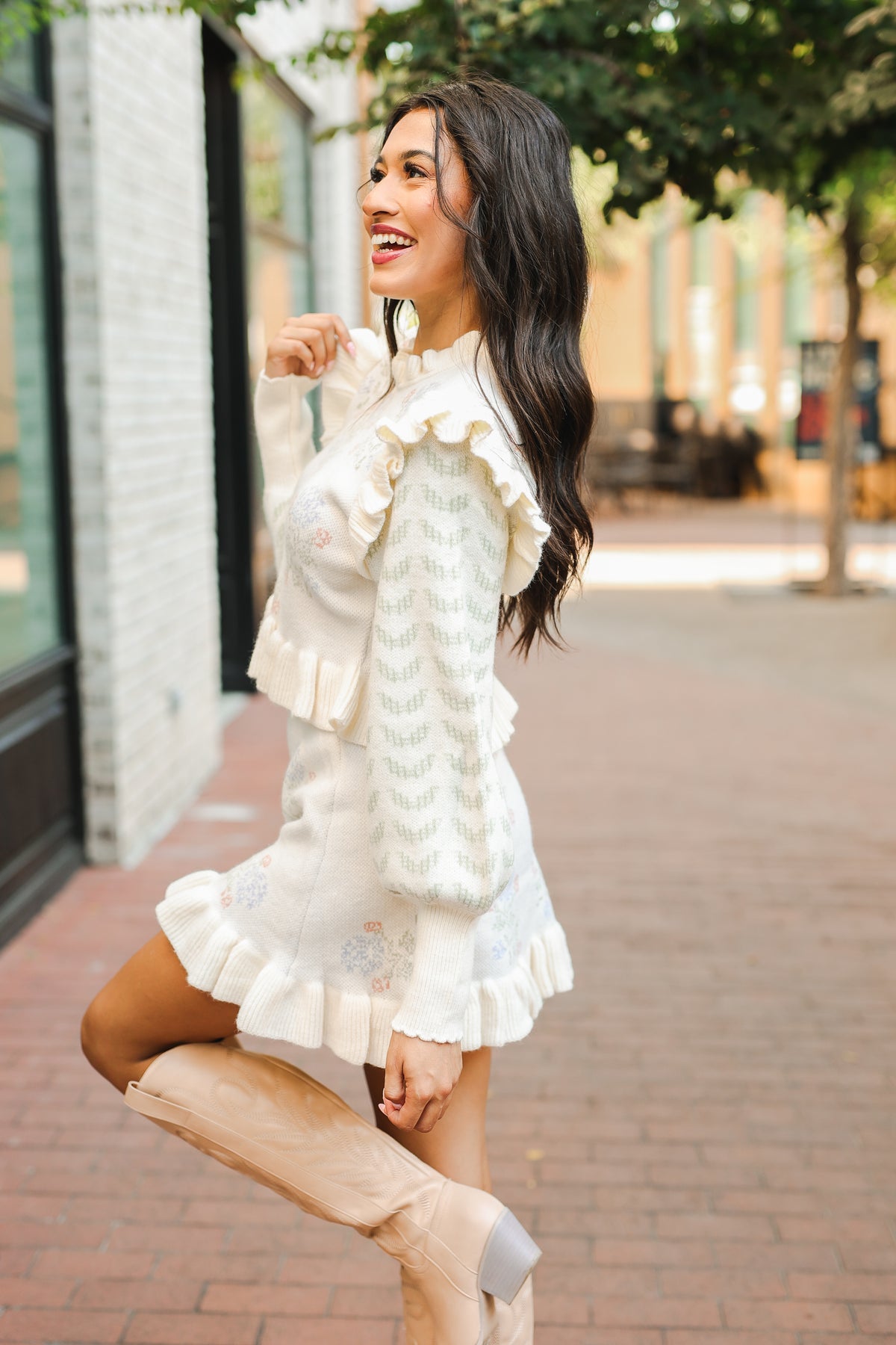 Woman in a white ruffled dress and beige boots standing on a brick sidewalk.
