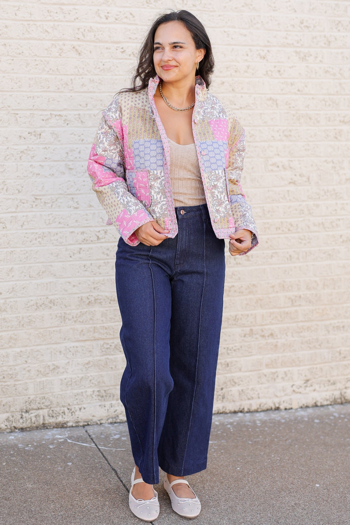 A woman stands outdoors against a light brick wall wearing a pastel patchwork quilted jacket with pink, blue, and beige panels, dark wide-leg jeans, a beige top, and light beige flats.
