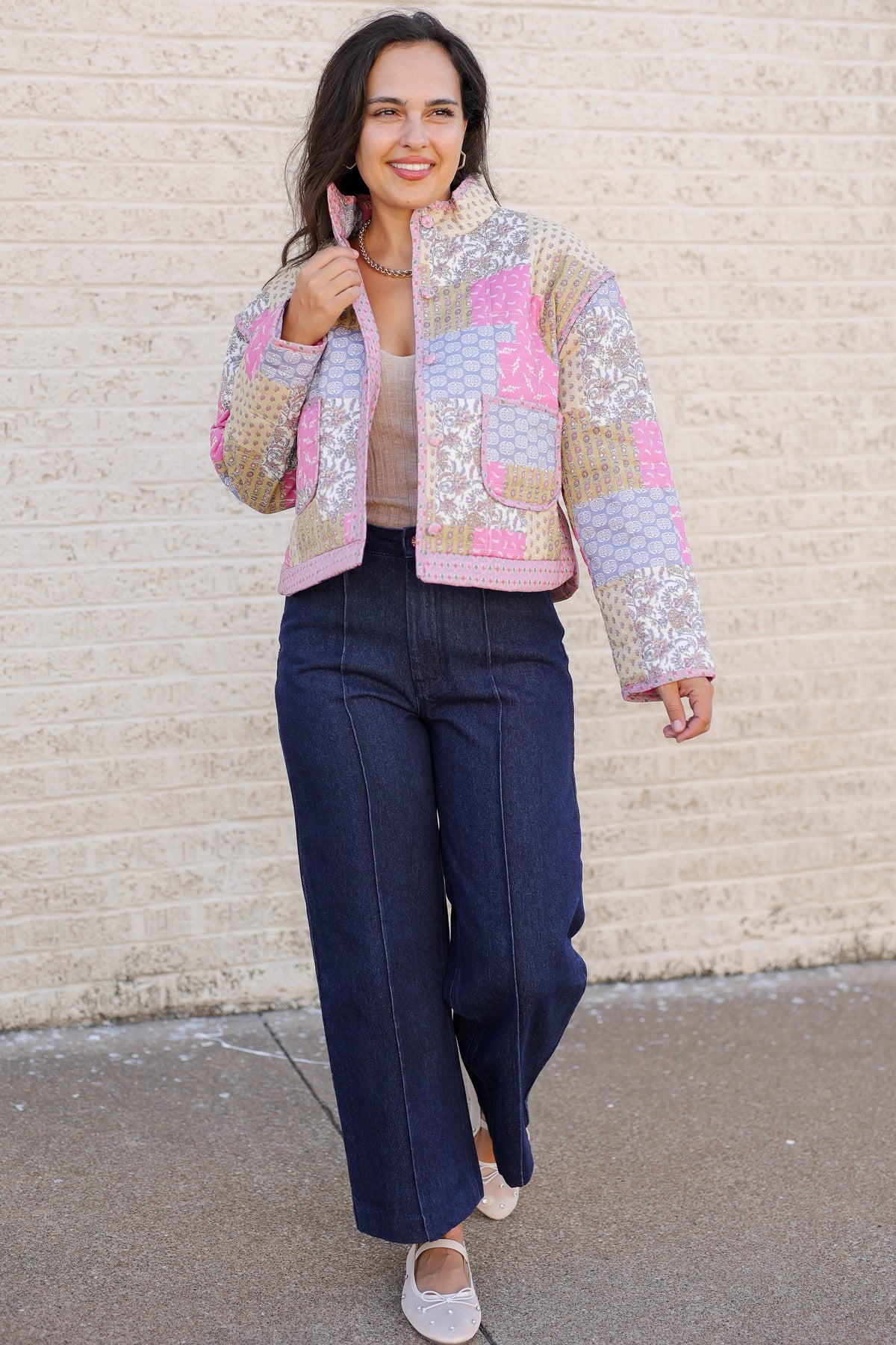 A woman wearing a colorful patchwork quilted jacket with pink, blue, and beige panels, paired with dark wide-leg jeans and white flats, standing against a light beige brick wall.