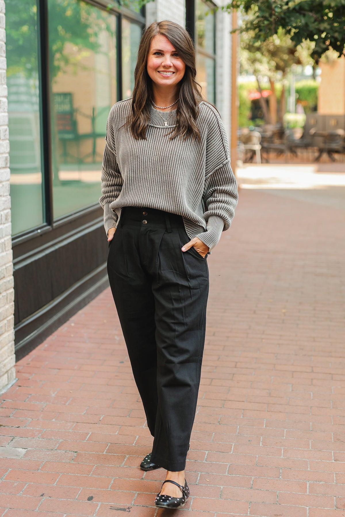 Woman wearing a gray sweater and black pants standing on a sidewalk.