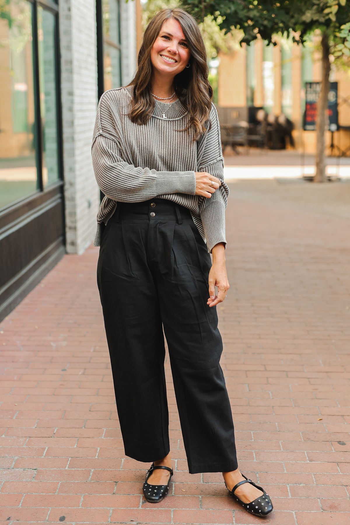 Woman wearing a gray sweater and black pants standing on a brick sidewalk.
