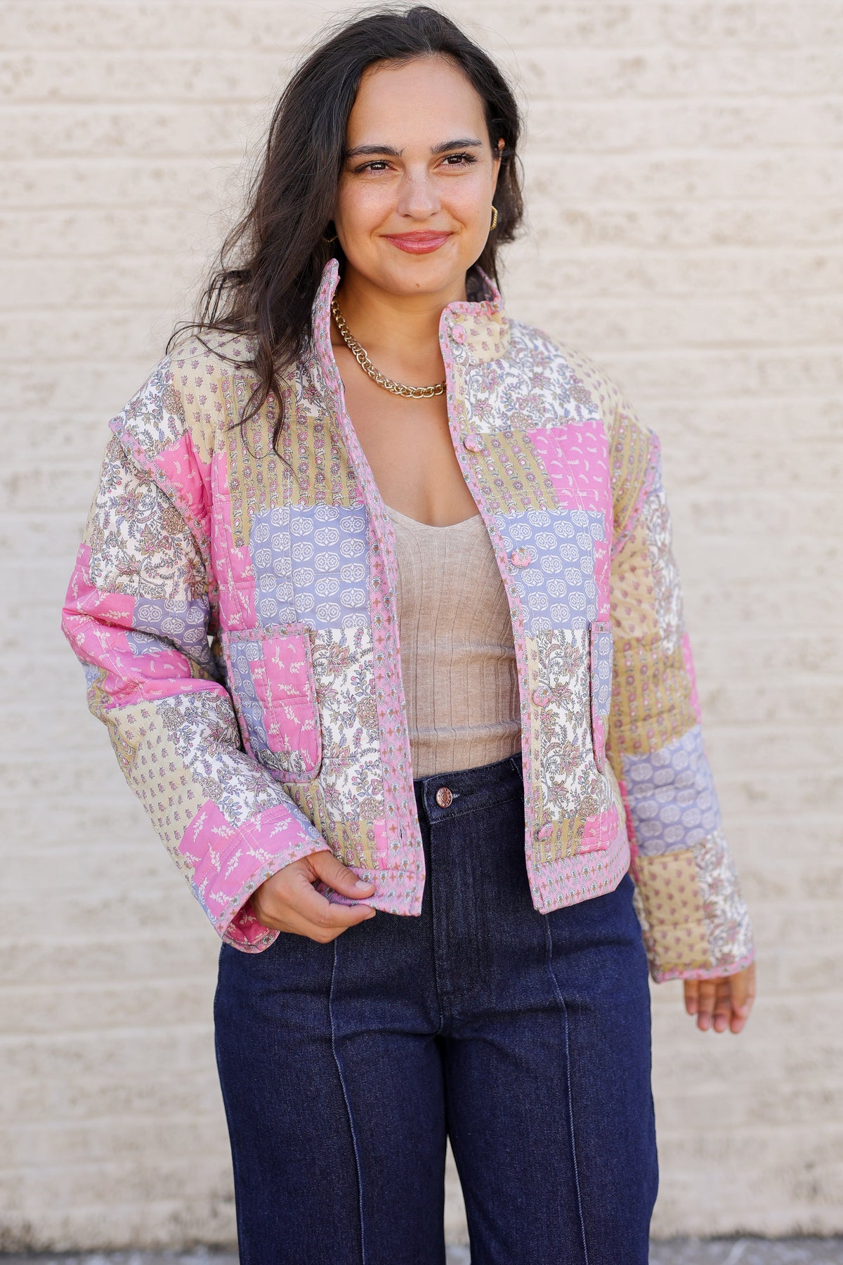 A woman wearing a colorful patchwork quilted jacket with pink, beige, and blue patterns, paired with a beige ribbed tank top and dark blue jeans, standing against a light beige brick wall.