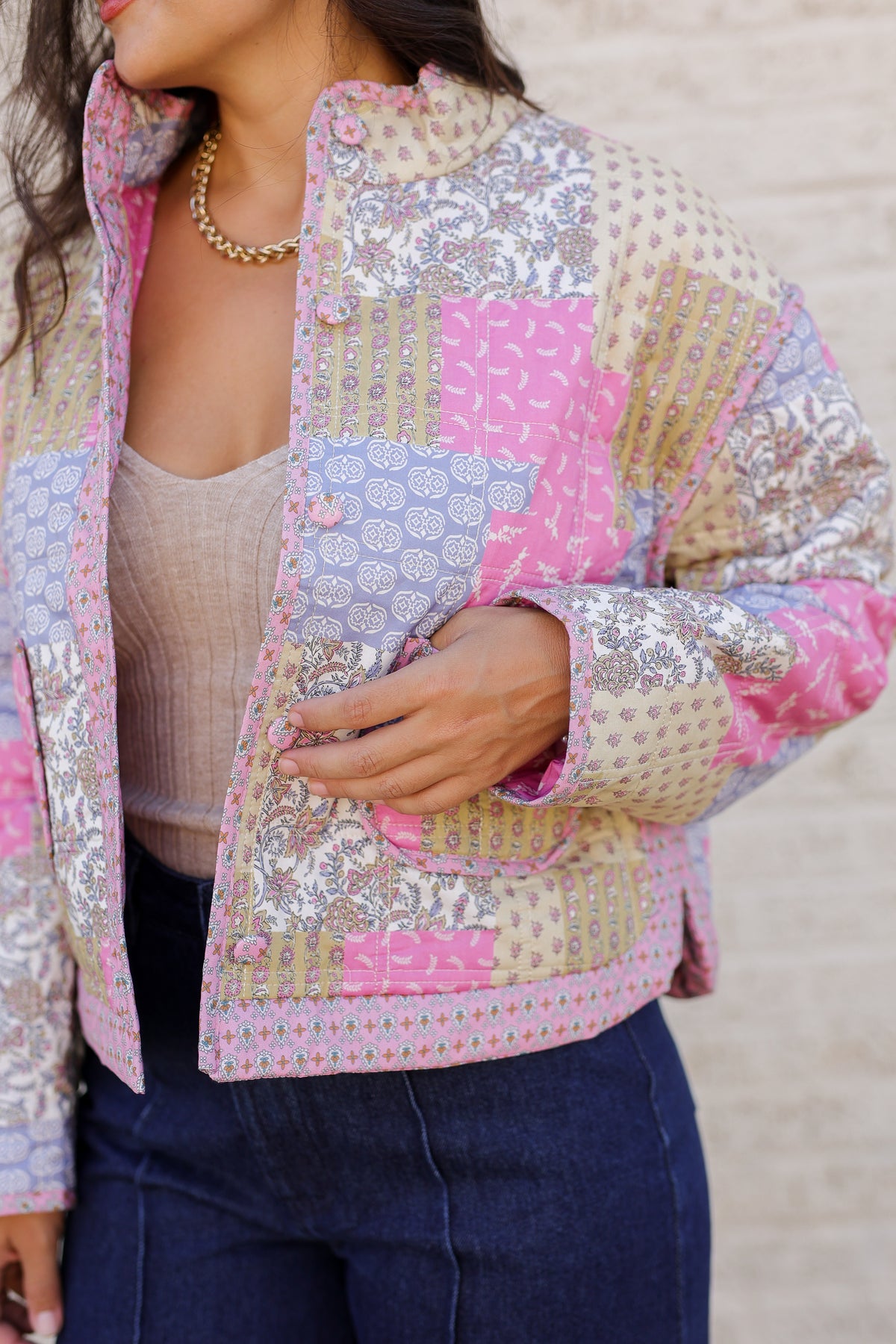A close-up of a woman wearing a pastel patchwork quilted jacket with pink, beige, and lavender panels, paired with a beige top and dark blue jeans.