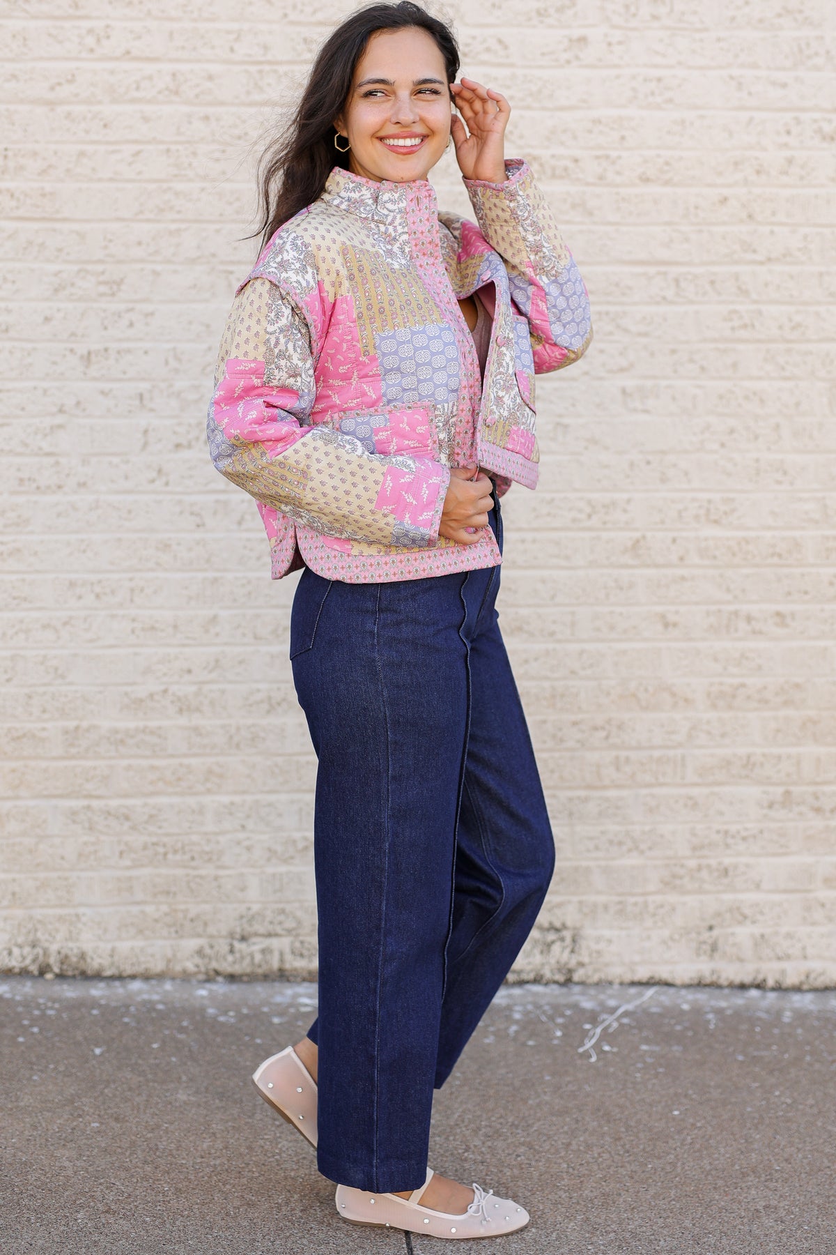 A woman stands outdoors against a light beige brick wall wearing a colorful patchwork quilted jacket with pink, purple, and gold tones, dark blue wide-leg jeans, and light pink flats.