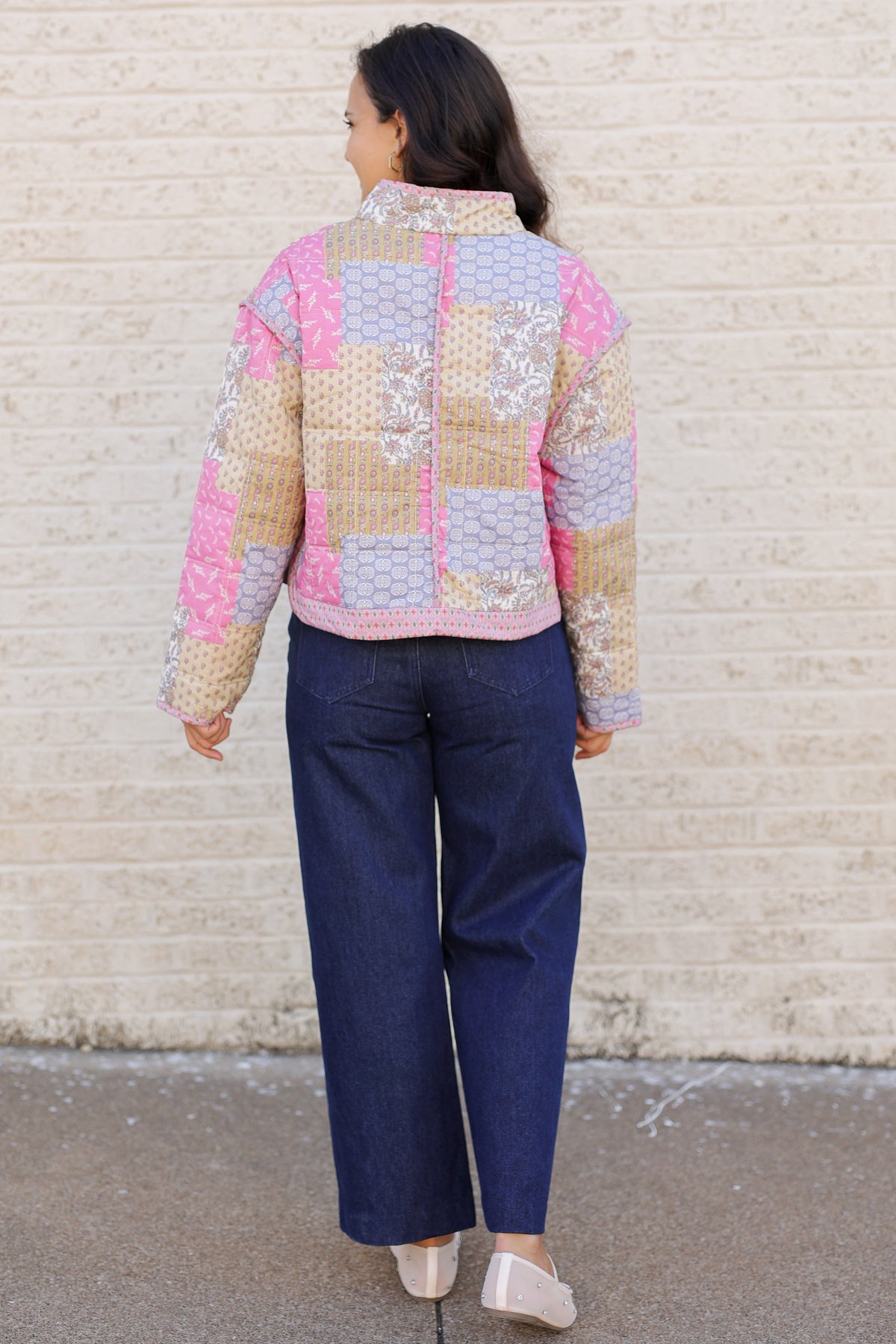 A woman viewed from behind wearing a pastel patchwork quilted jacket with pink, beige, and lavender panels, paired with dark blue jeans and white flats, standing against a light brick wall.